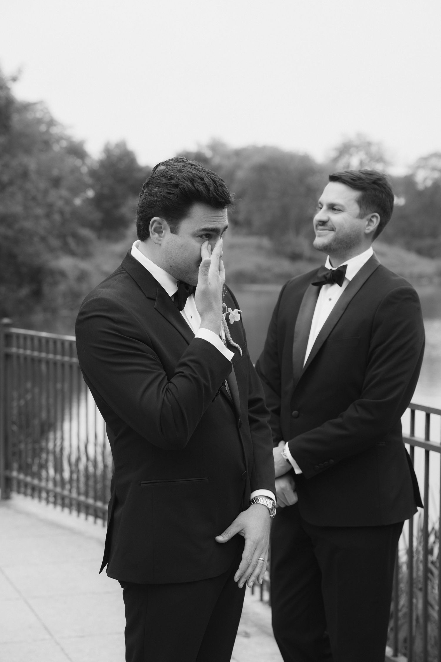 Black and white photo of two grooms in tuxedos sharing an emotional first look moment on an outdoor terrace.