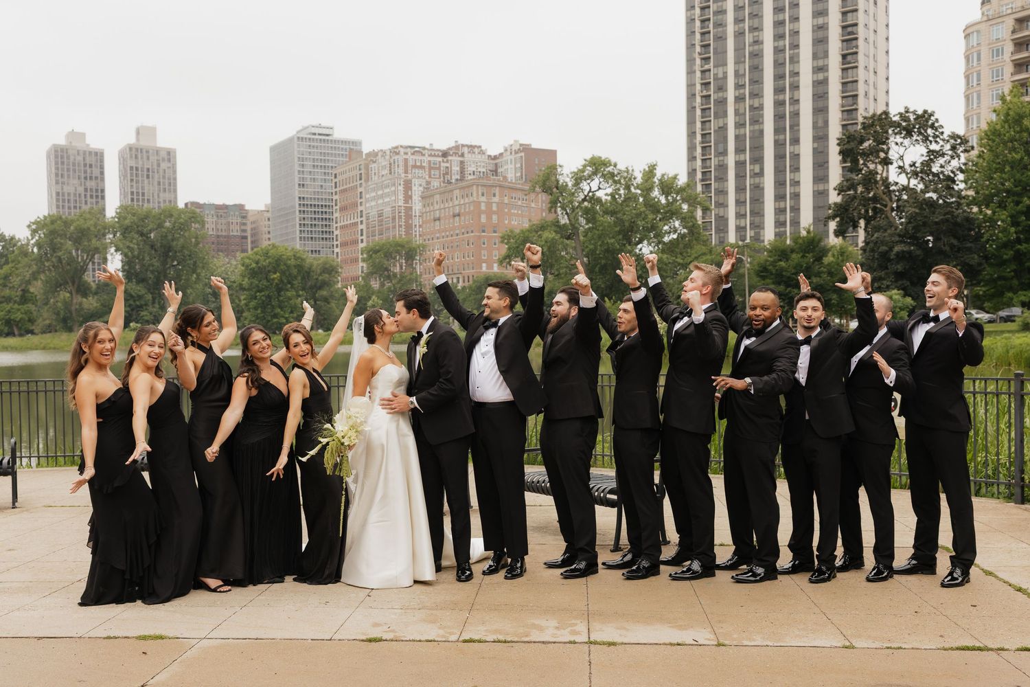 Wedding party in black attire cheering outdoors with Chicago skyline and lush greenery in the background.