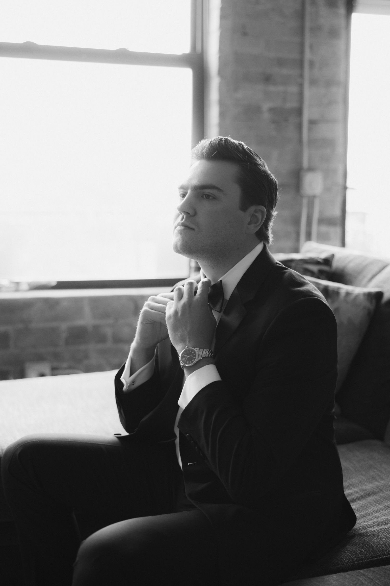 Black and white photo of a groom in a tuxedo adjusting his bow tie while seated near a window.