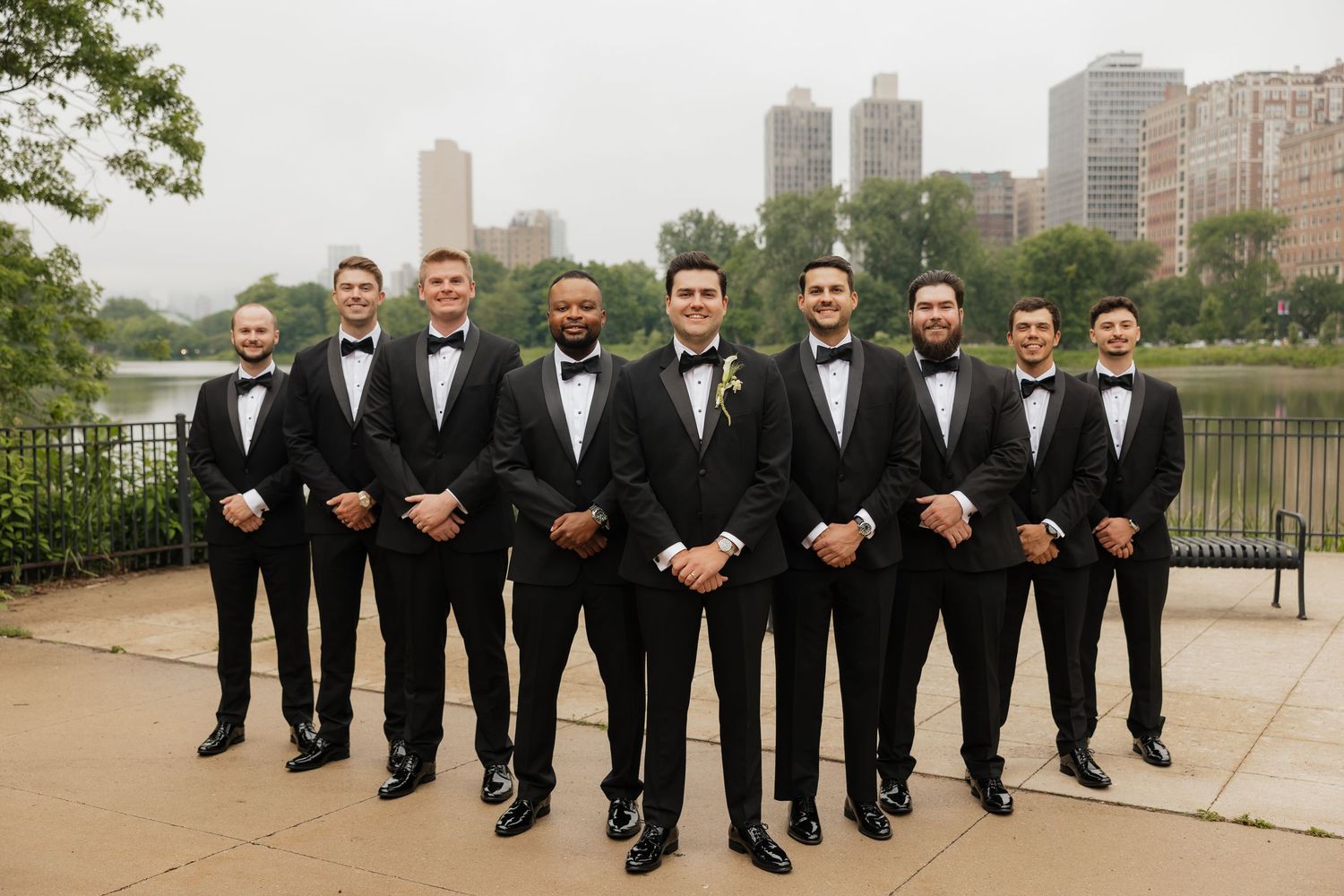 Groom and groomsmen in black tuxedos with bow ties pose outdoors near a river with city skyline backdrop.