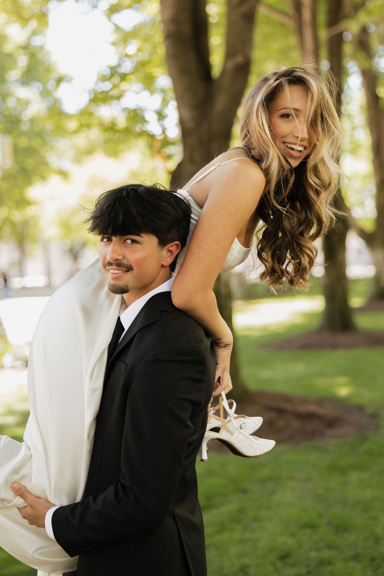 Groom in black suit carries bride in white dress on his back outdoors in a park setting.