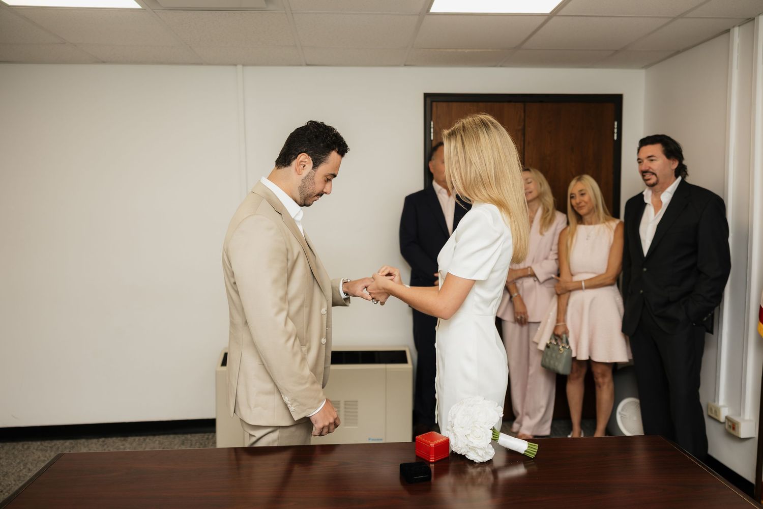 Couple exchanging rings during an intimate courthouse wedding ceremony with smiling guests watching in background.