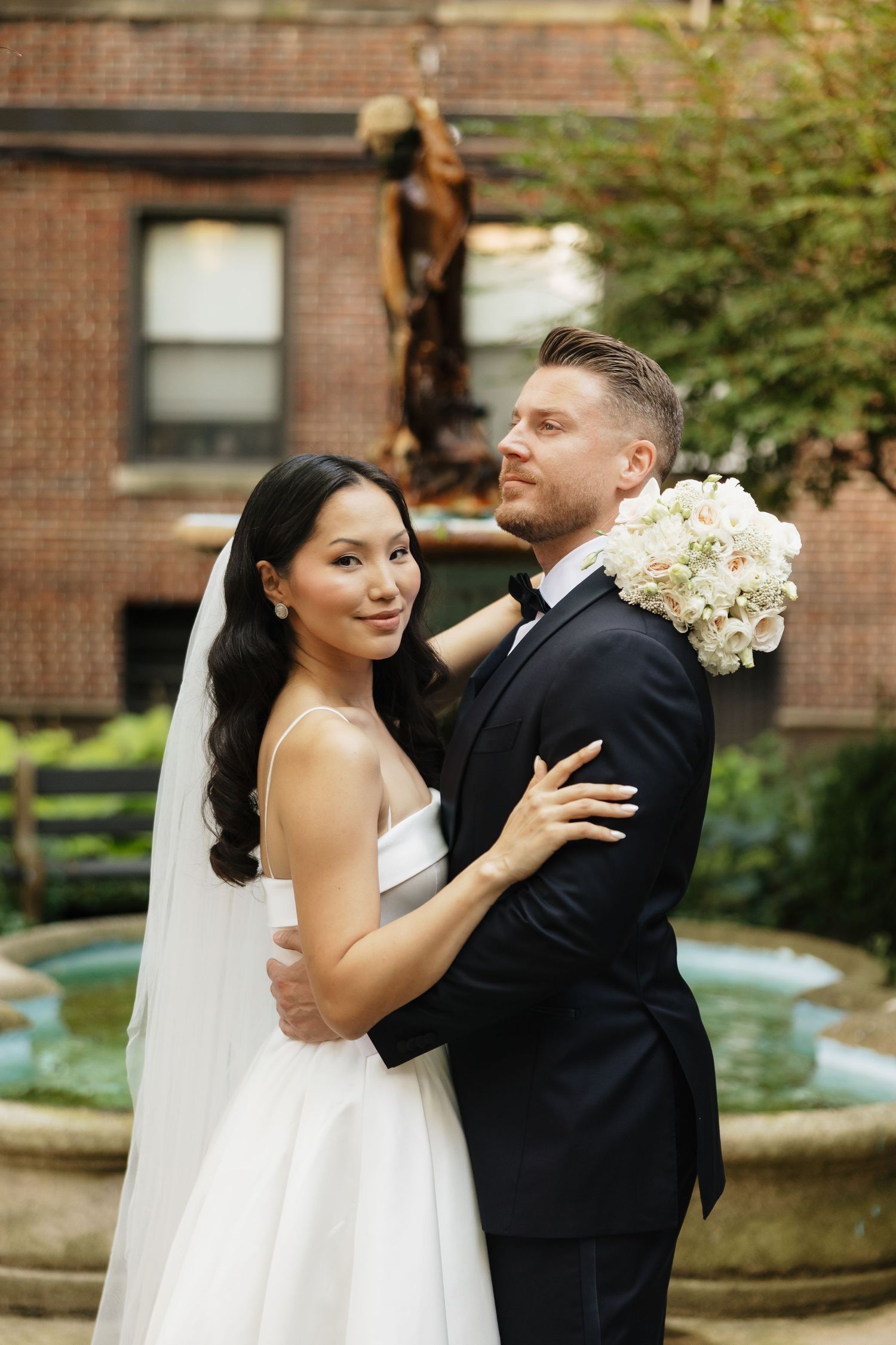 Bride and groom embracing in a garden courtyard with fountain, bride holding white floral bouquet in wedding attire.