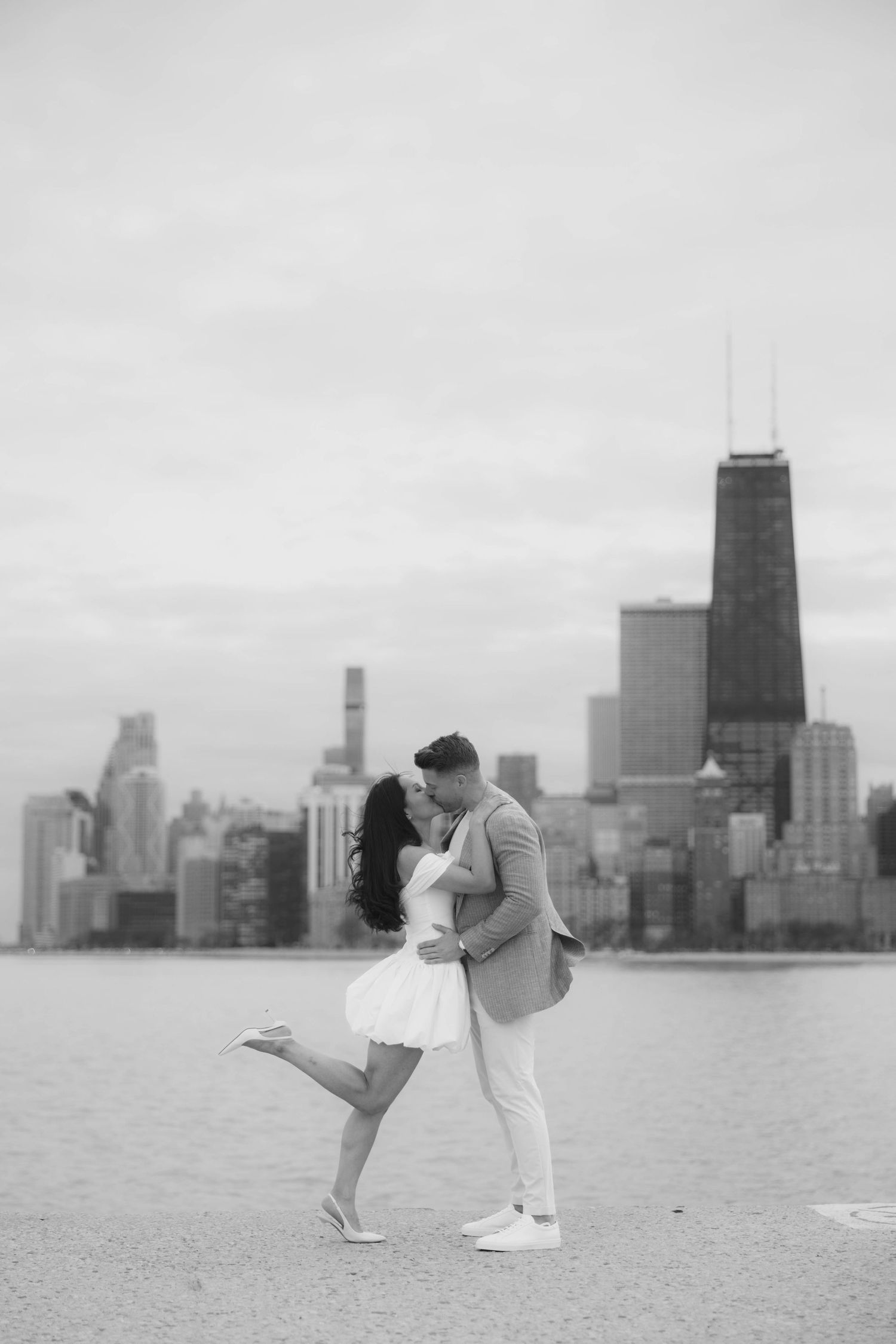 Romantic black and white photo of a couple kissing with Chicago skyline, woman lifting her leg playfully.