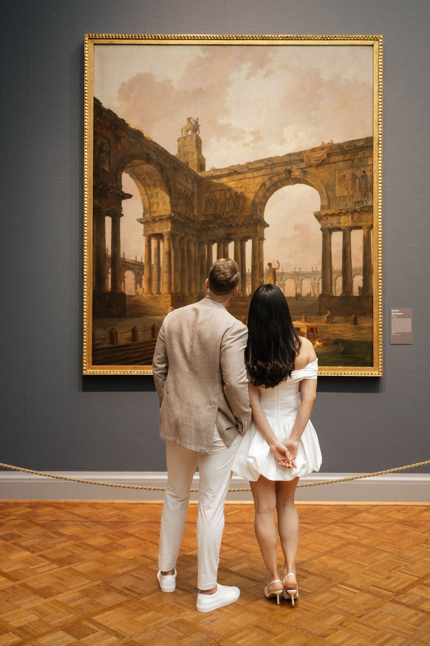 Couple in white attire viewing a classical ruins painting in an elegant art museum gallery.
