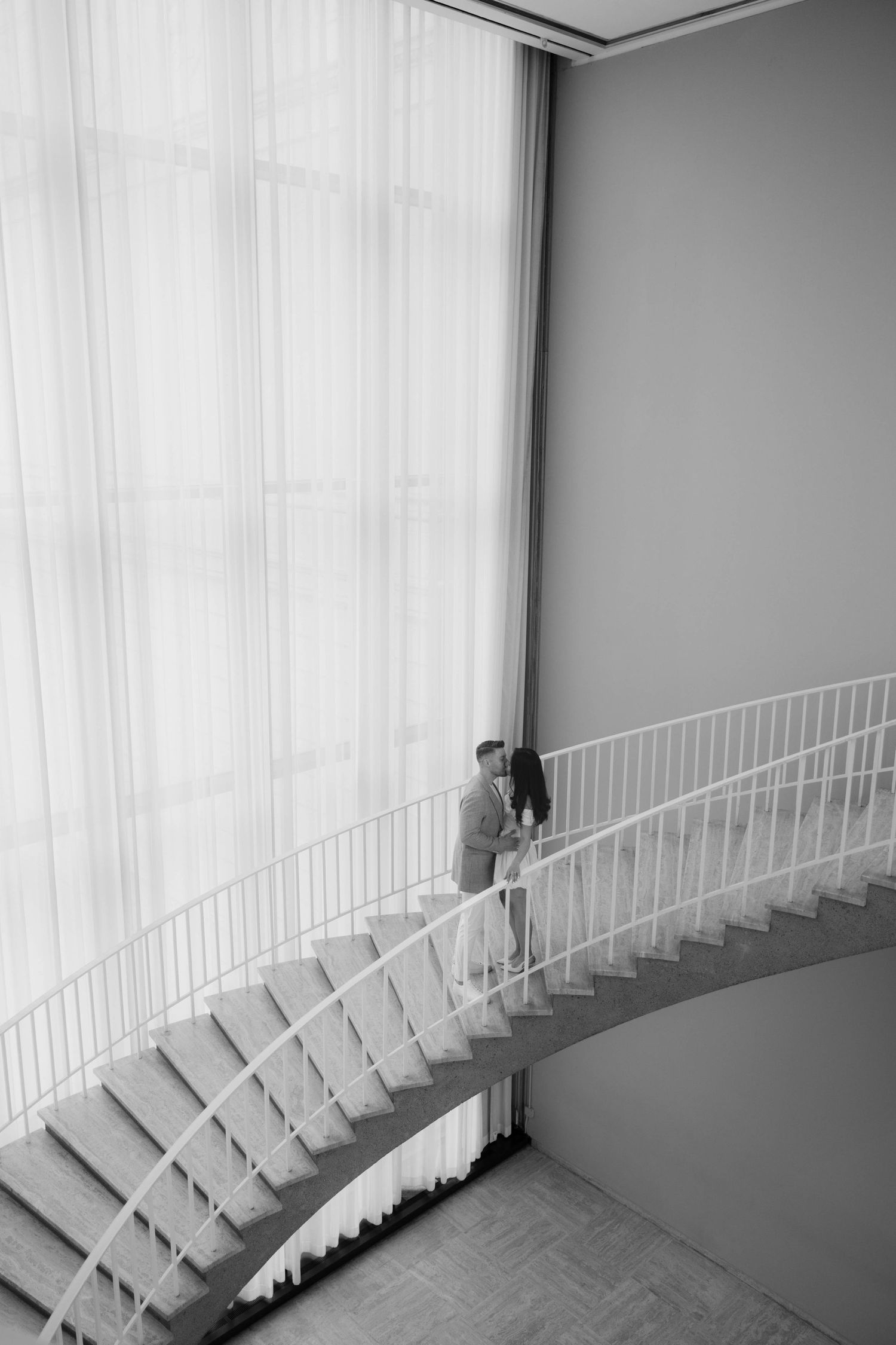 Black and white photo of a couple standing on a curved staircase near sheer curtains in an elegant interior.
