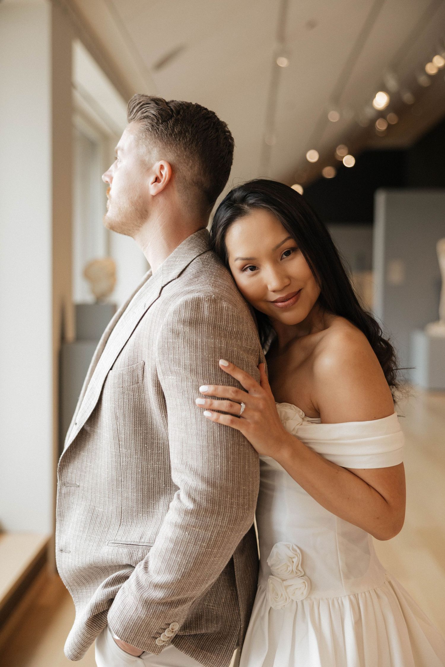Couple poses romantically in museum gallery, bride in white dress leaning on groom's shoulder.