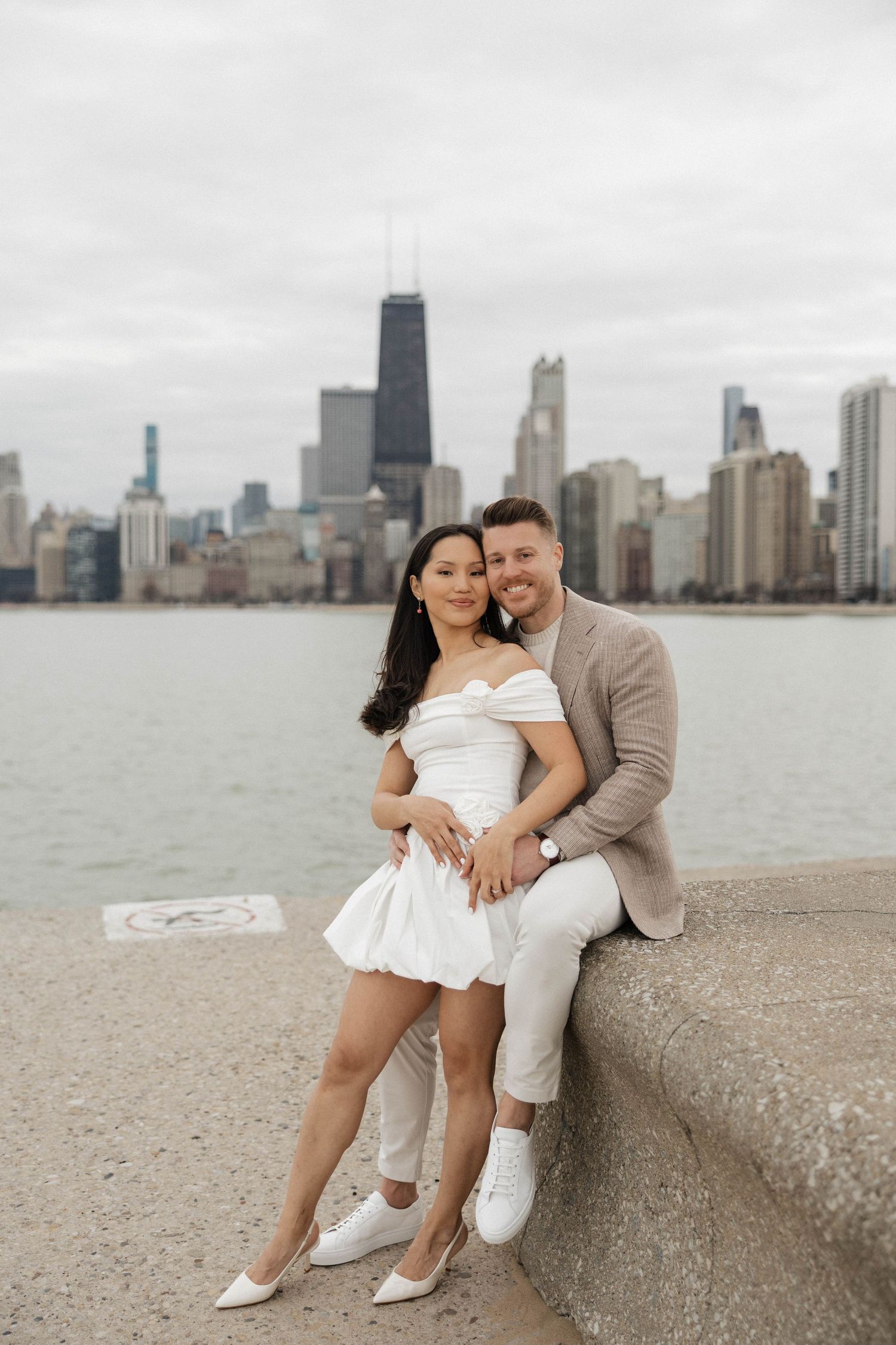 Couple poses by Lake Michigan with Chicago skyline backdrop, woman in white dress, man in beige suit.