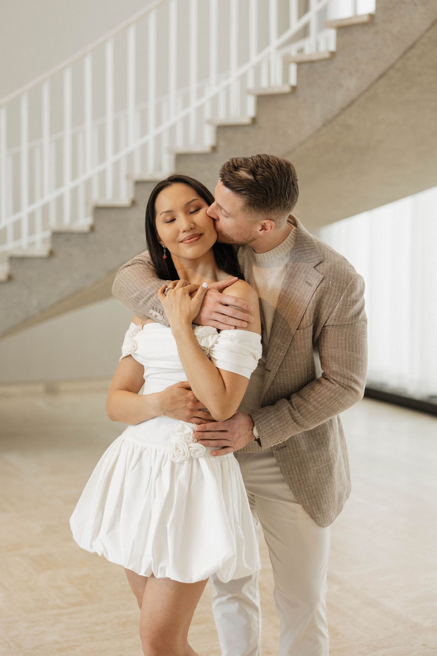 A couple embraces near a white spiral staircase, man in beige suit kisses woman in white mini dress.