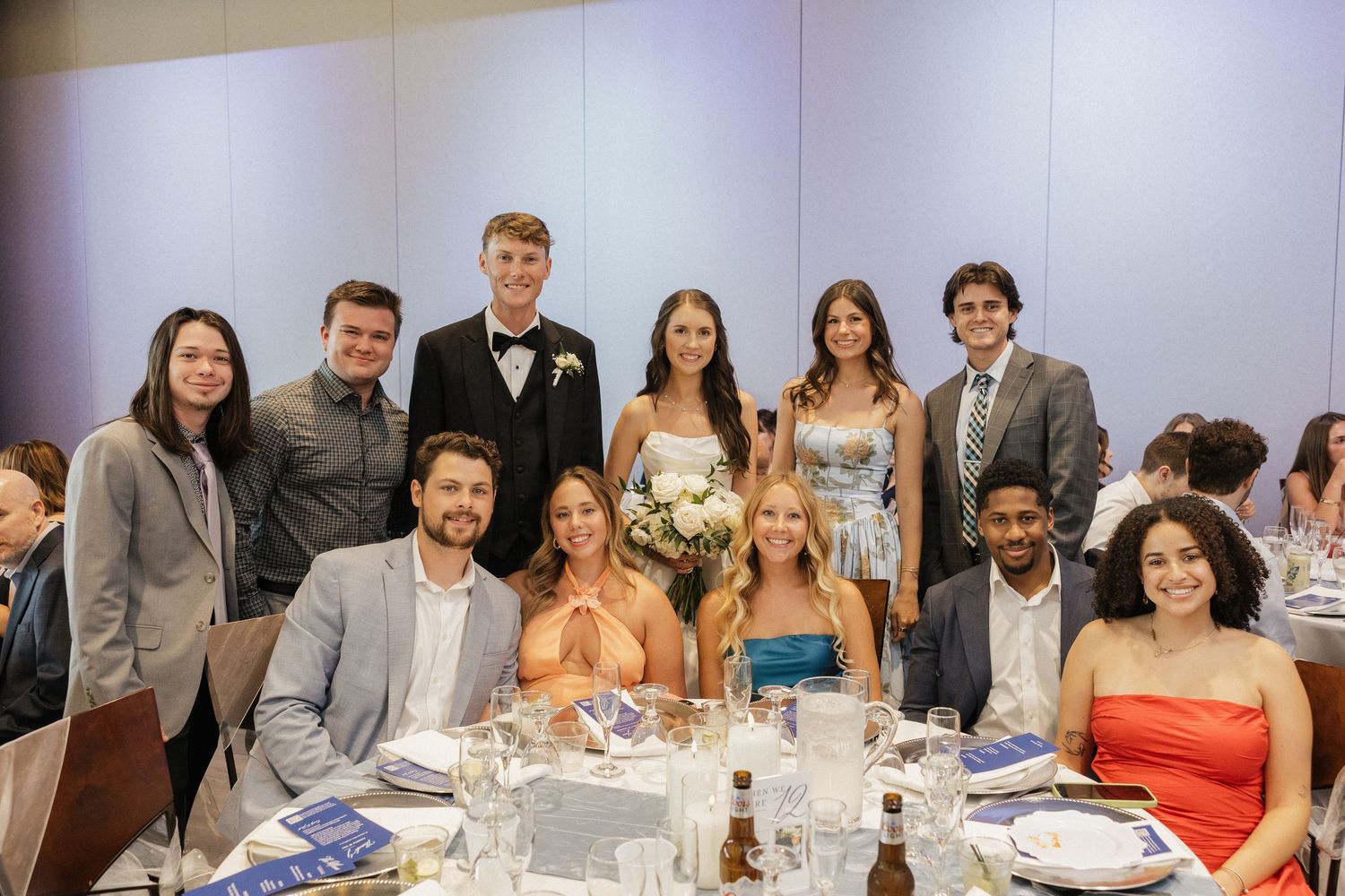 Group of elegantly dressed young adults posing together at a wedding reception dinner table with floral centerpiece.