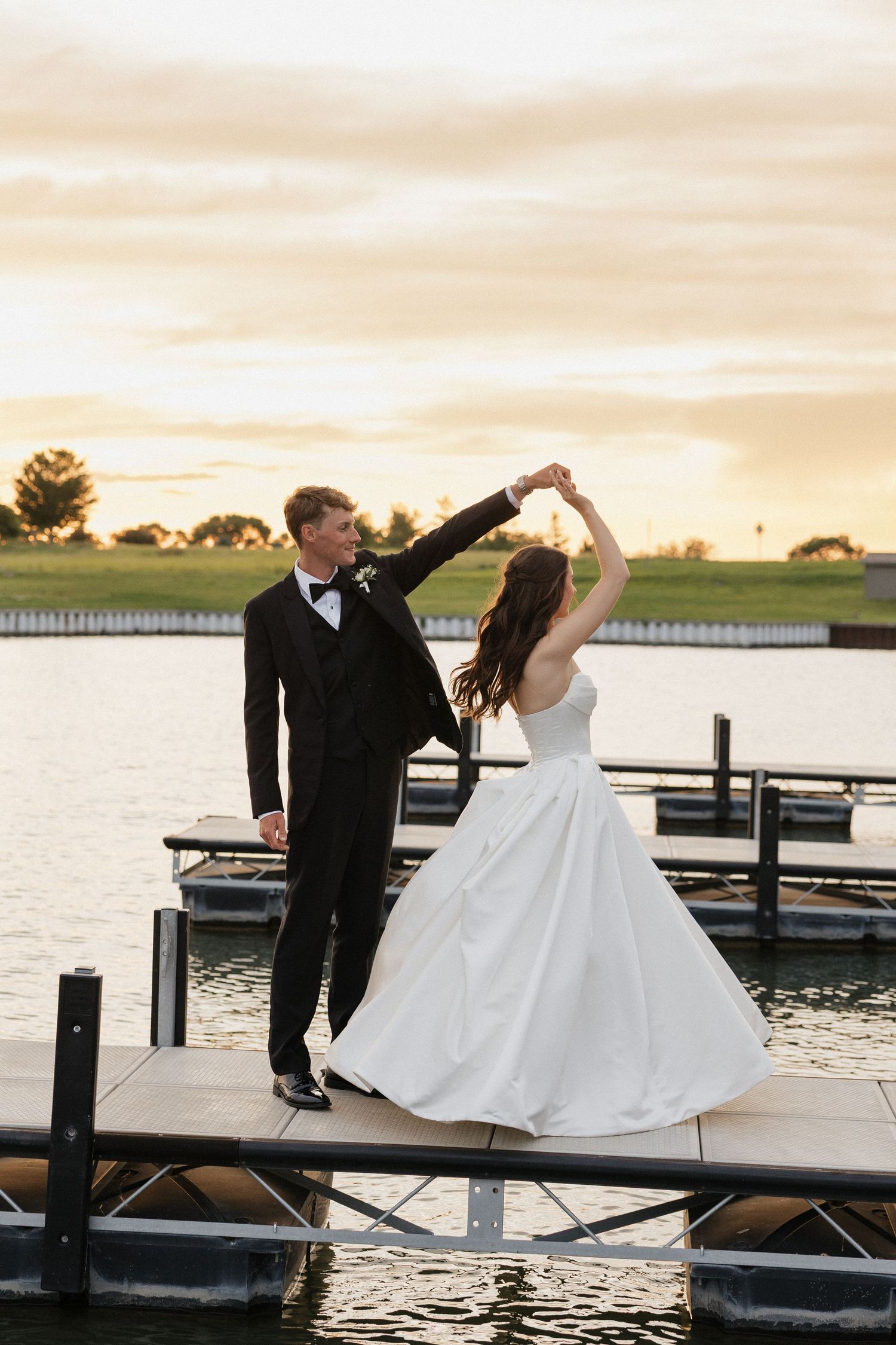 Bride and groom dancing on a dock at sunset, with the groom spinning his bride in a white ballgown by the water.