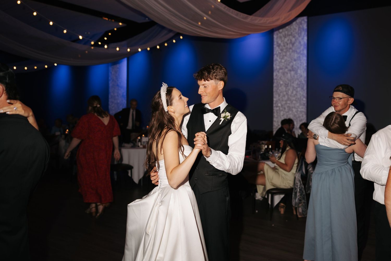 Bride and groom share their first dance at a wedding reception with blue lighting and string lights.