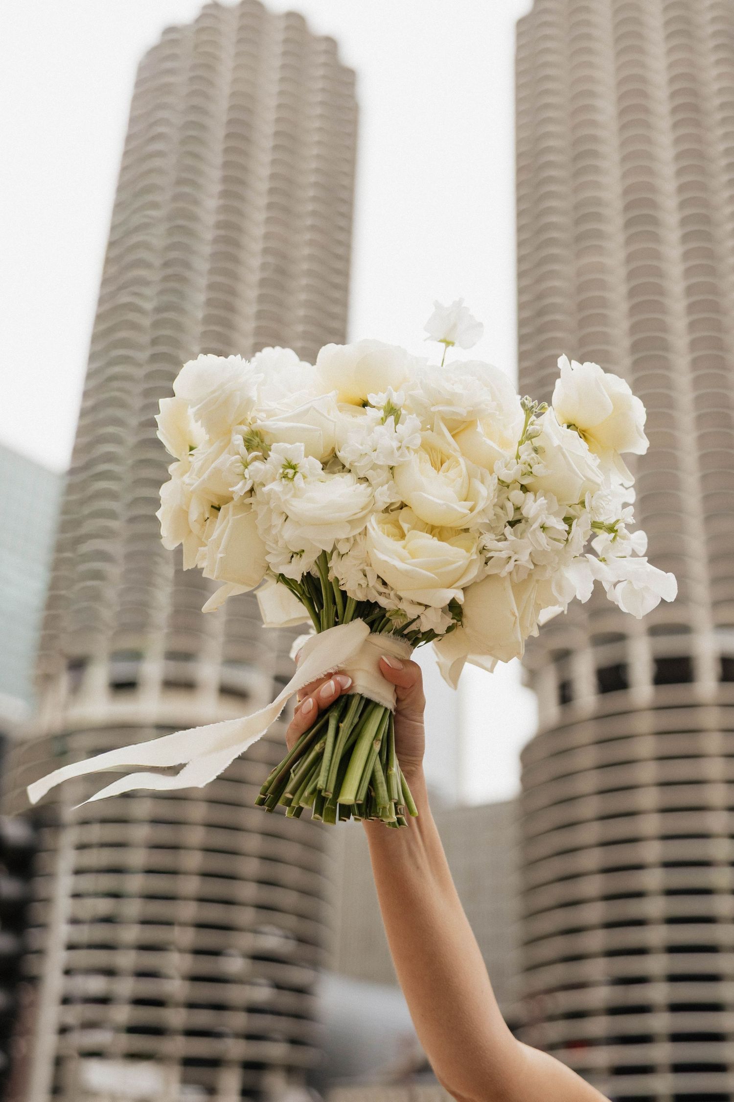 A hand holds a white wedding bouquet of ranunculus and roses with ribbon, against urban skyscrapers backdrop.