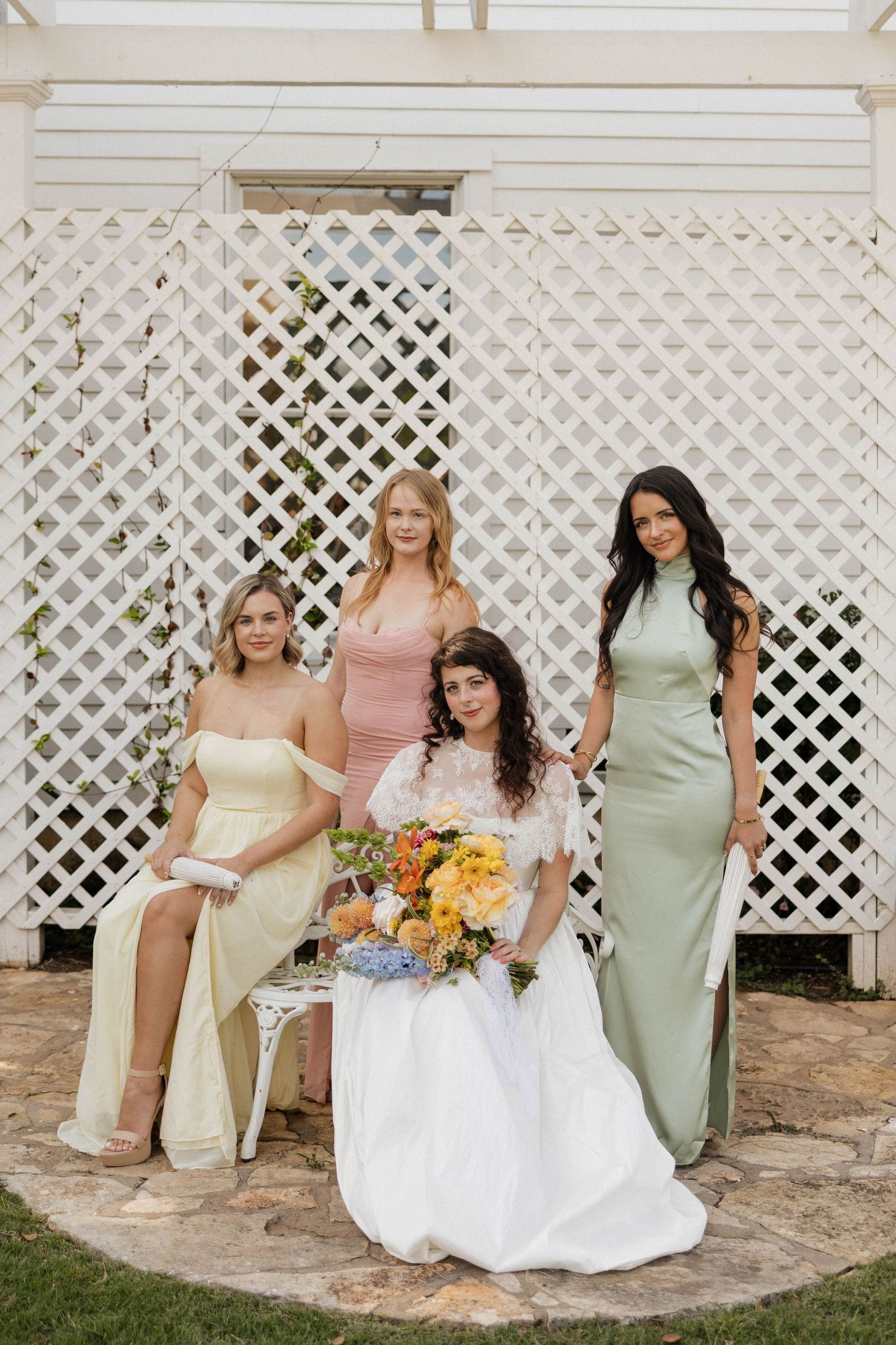 Bride in white gown holds colorful bouquet with three bridesmaids in pastel dresses posed before white lattice backdrop.