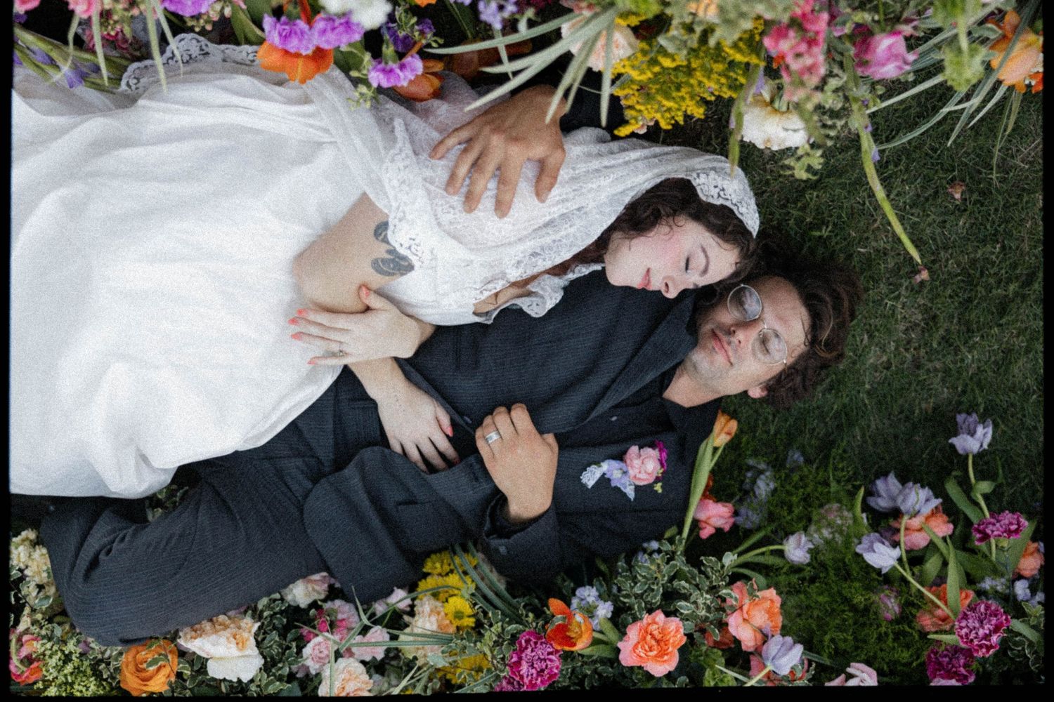 Bride and groom lying among colorful flowers on grass, wearing wedding attire, viewed from above.