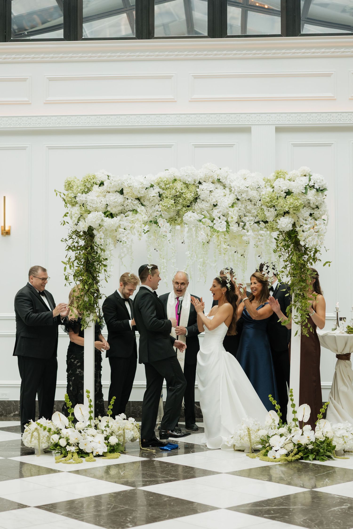 Bride and groom share a joyful moment under a floral chuppah adorned with white flowers and greenery during indoor wedding ceremony.