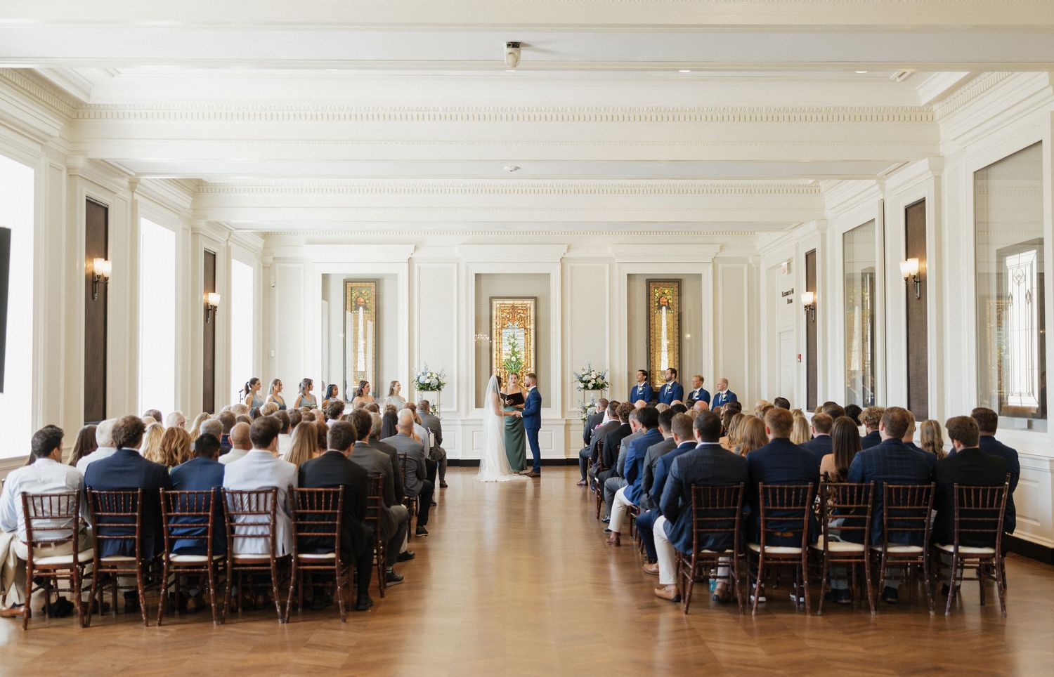 A panoramic view of a formal dining event in an elegant white-walled room with wooden chairs and large windows.