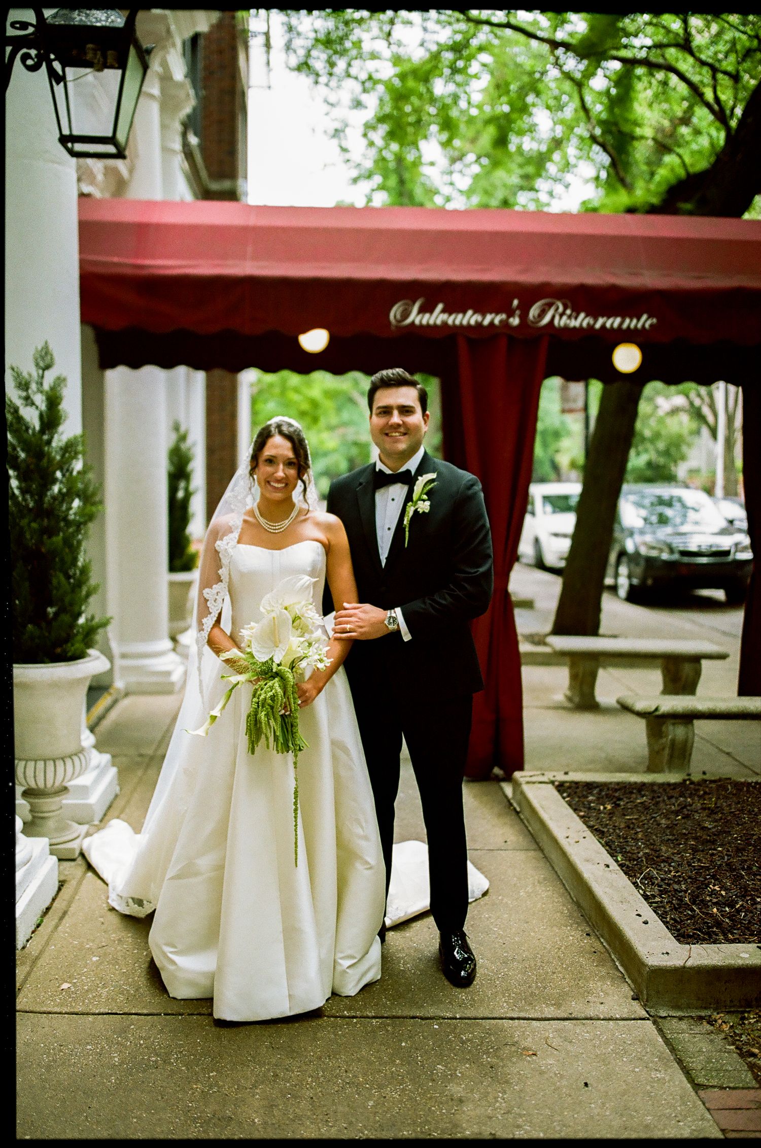 Bride and groom pose outside Salvatore's Ristorante, bride in white gown holding greenery bouquet, groom in black tuxedo.