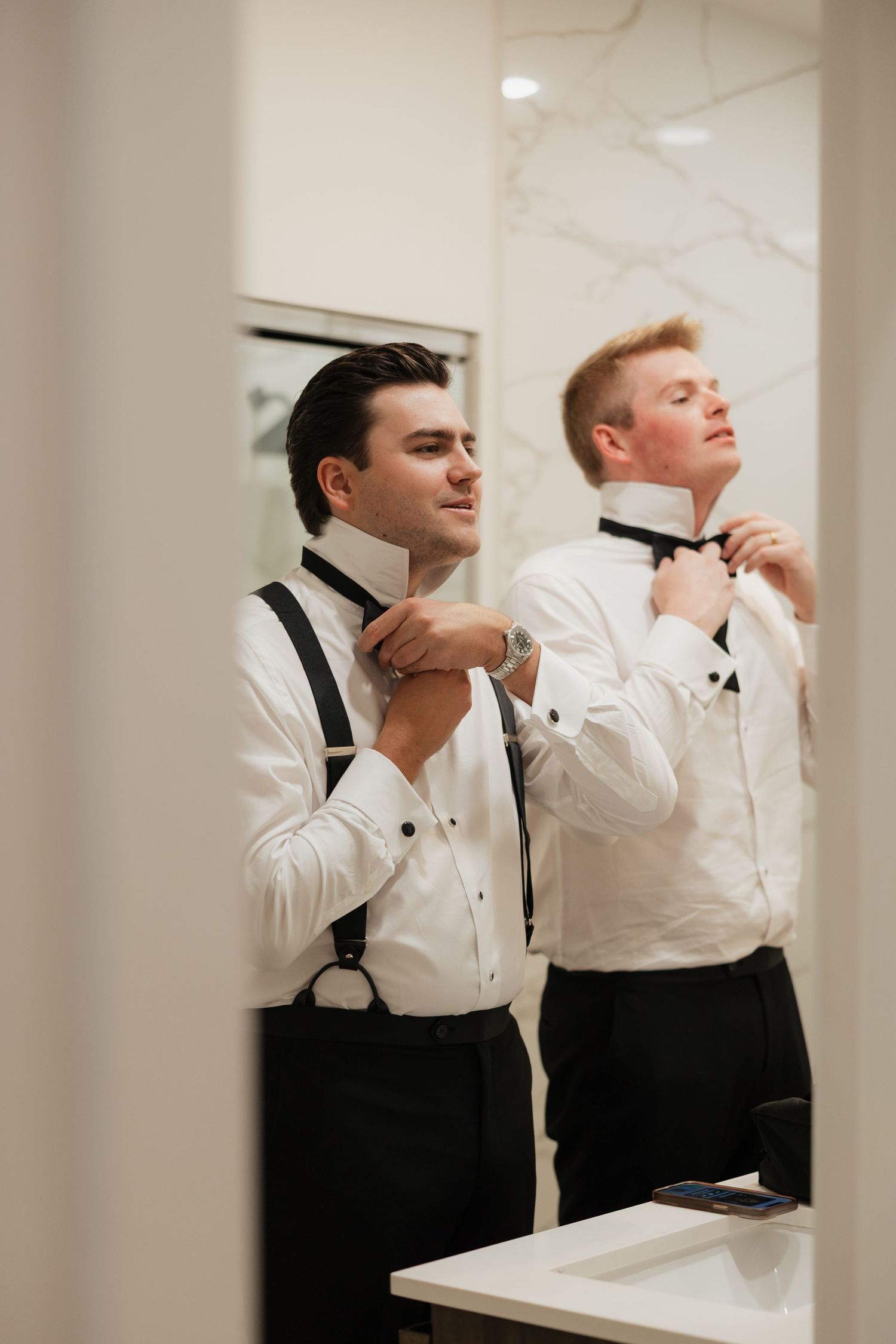 Two groomsmen in white shirts and suspenders adjusting black bow ties before a wedding ceremony.