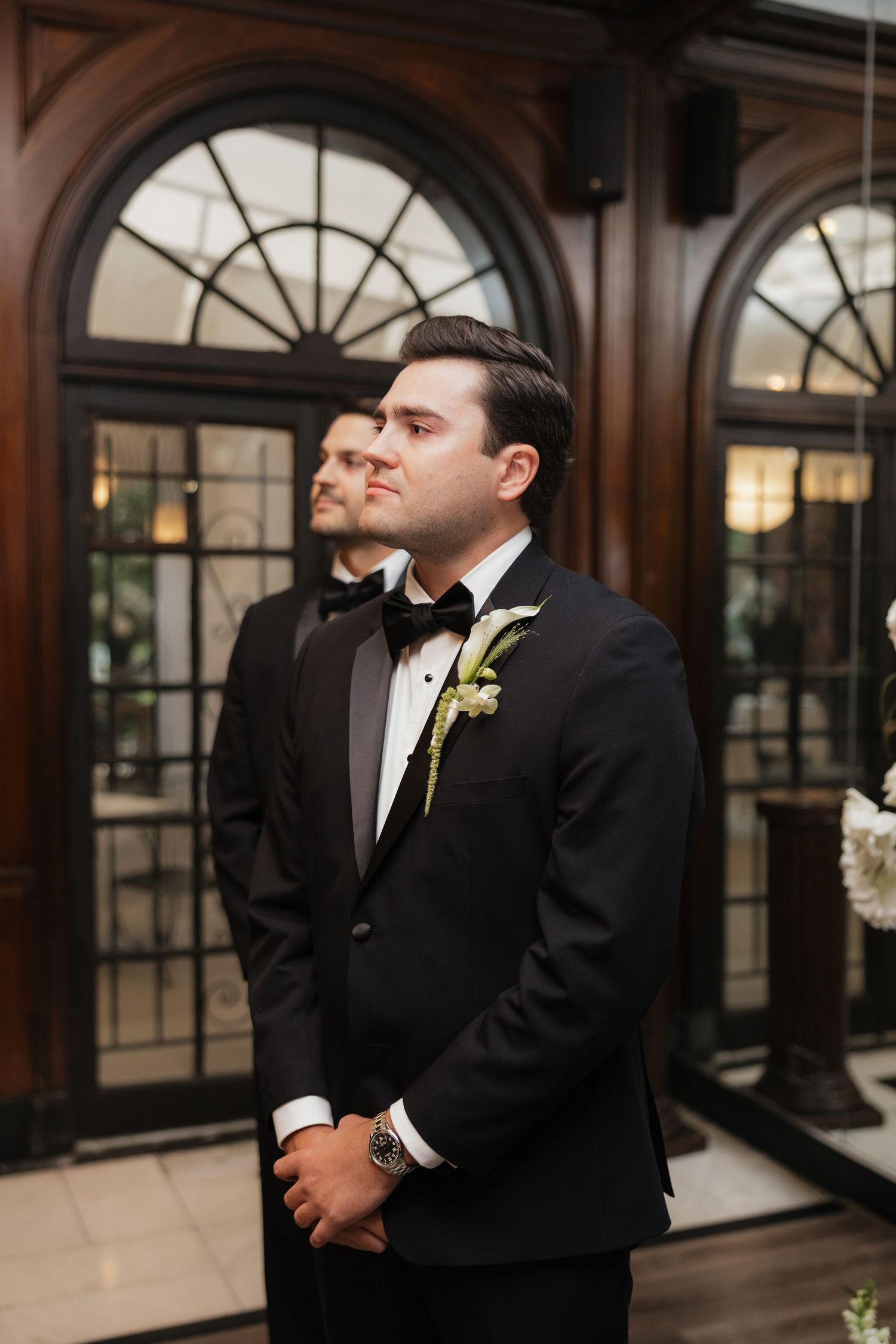 Groom in black tuxedo with floral boutonniere stands at wedding altar, groomsman visible in background.