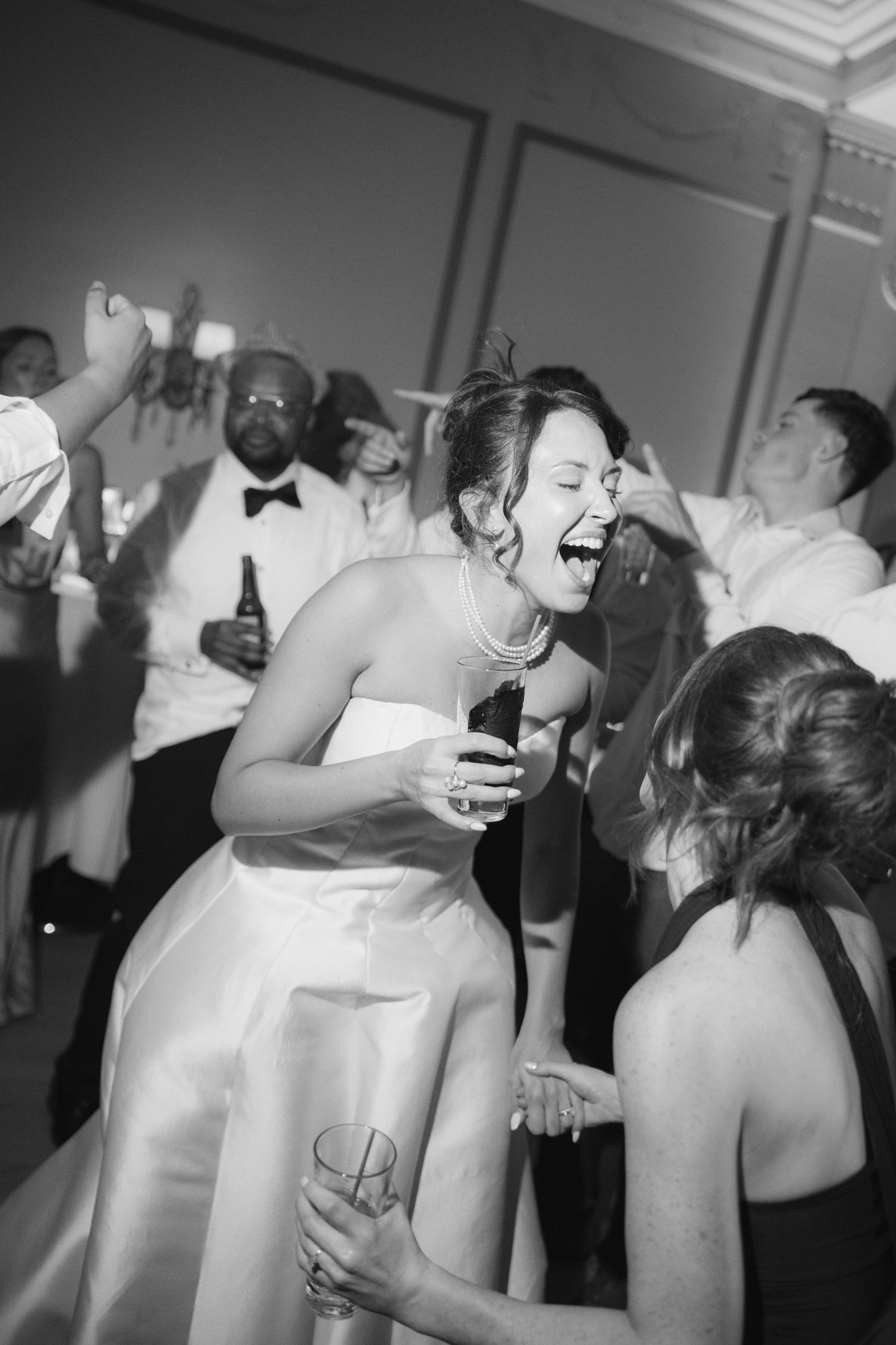 Black and white photo of a laughing bride dancing joyfully with guests at her wedding reception.