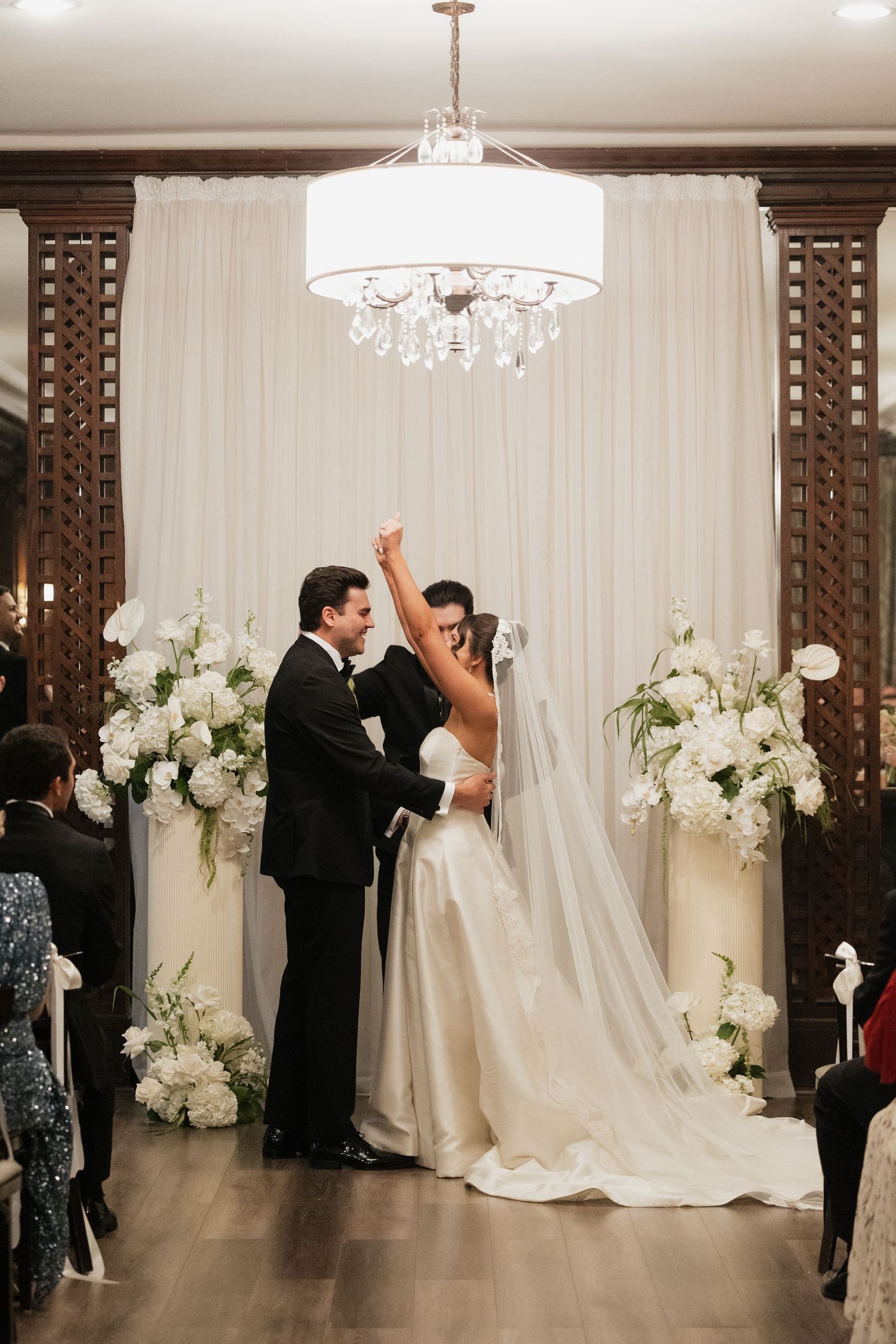 Bride and groom celebrate at indoor wedding ceremony with white floral arrangements and elegant draped backdrop.