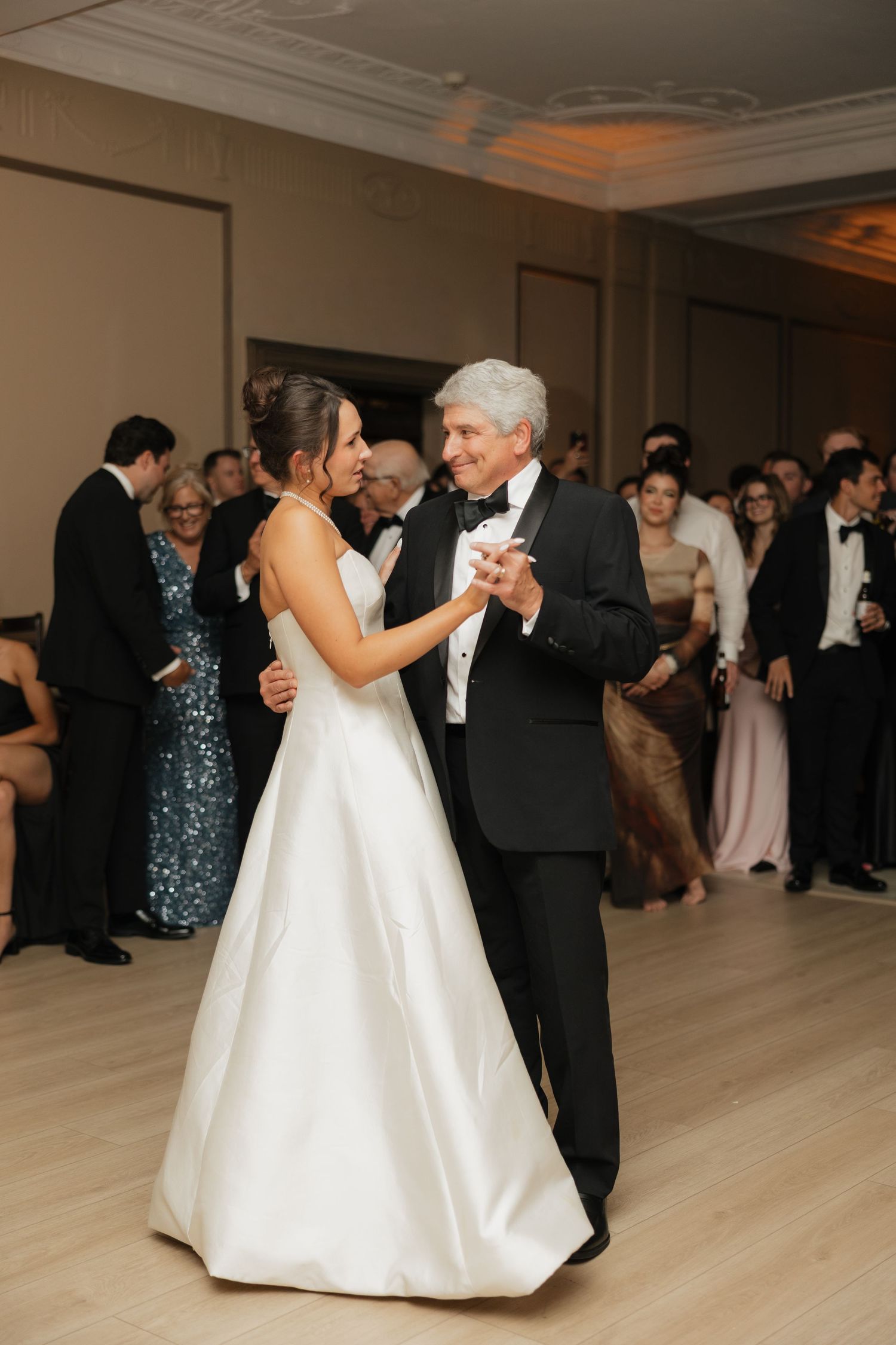 Bride in white gown shares father-daughter dance at elegant wedding reception with guests watching.