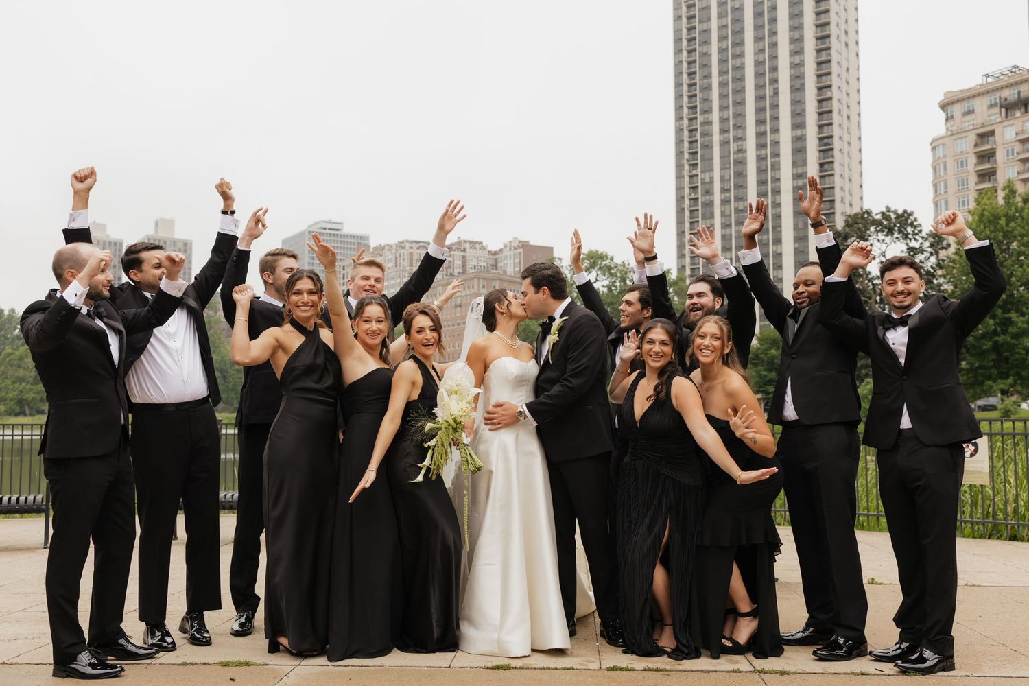 Wedding party in black attire cheers around kissing bride and groom outdoors with city skyline backdrop.