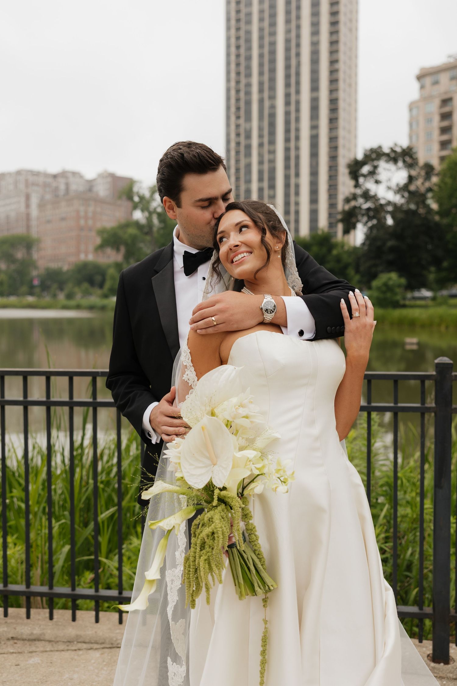 Bride and groom embrace outdoors near a pond, groom kissing bride's cheek, bride holding white calla lily bouquet.