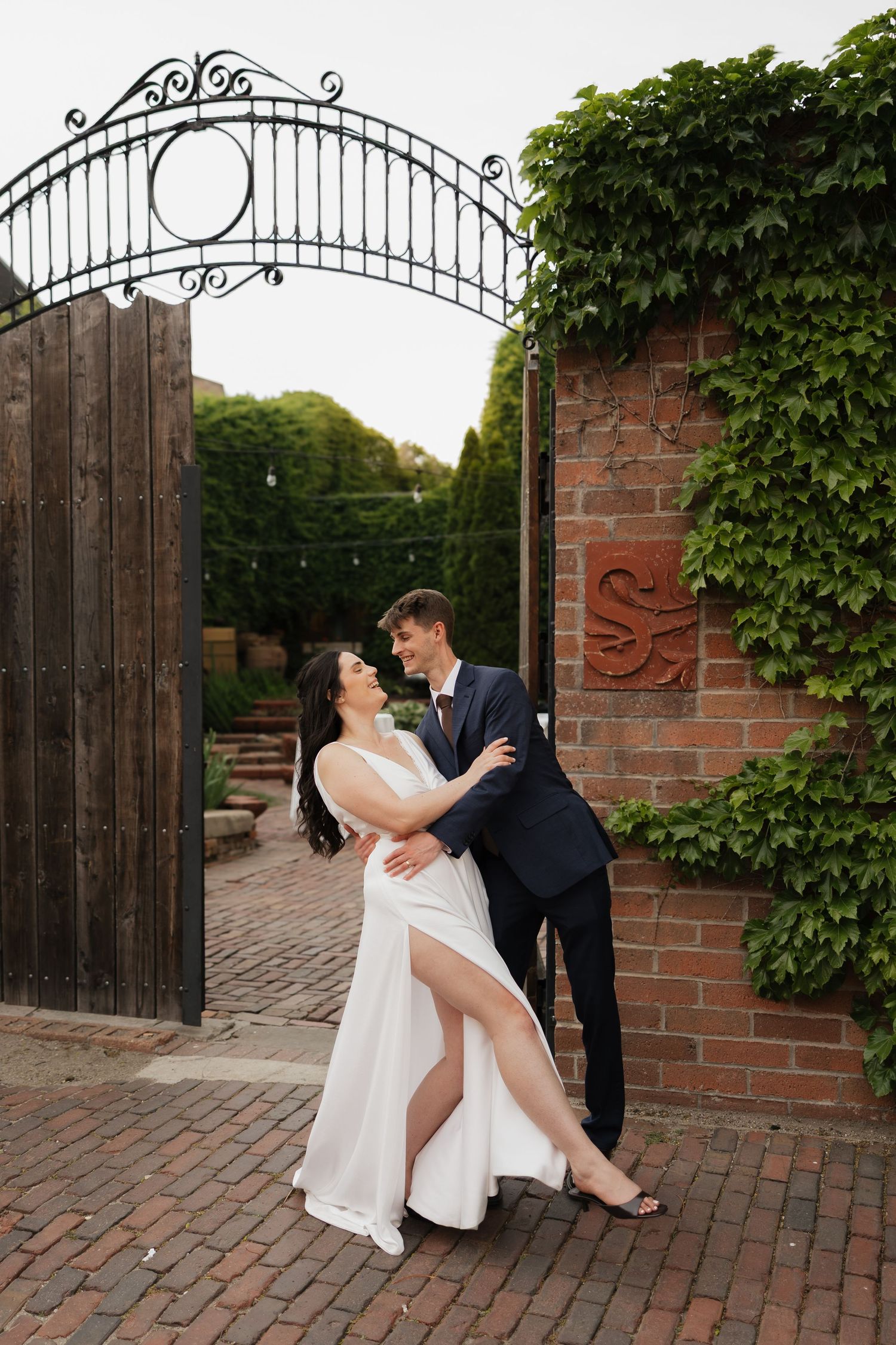Bride and groom embrace romantically by an ivy-covered brick wall and ornate iron gate on their wedding day.