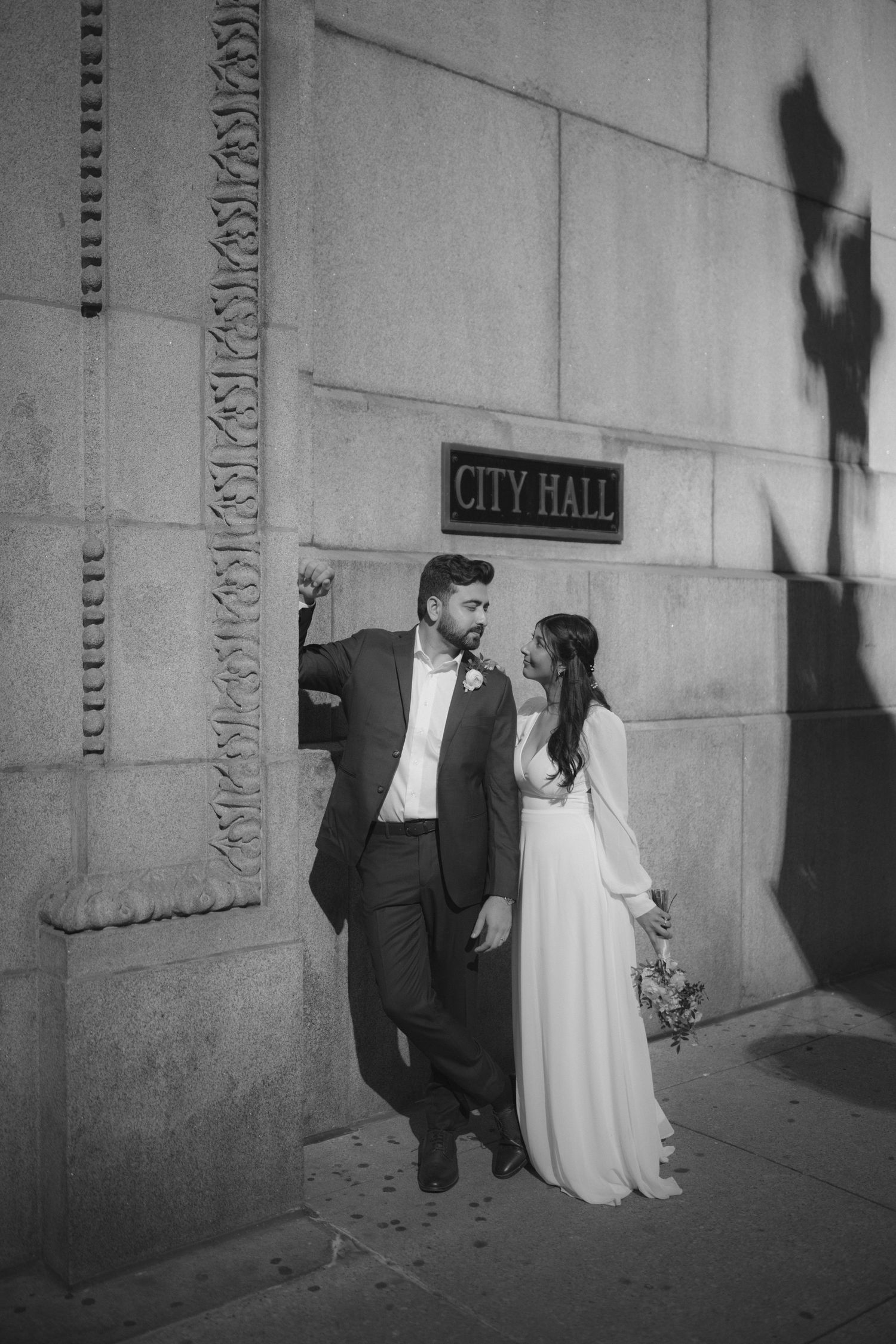 A romantic black and white wedding photo of a couple posing by a City Hall sign on a building wall.