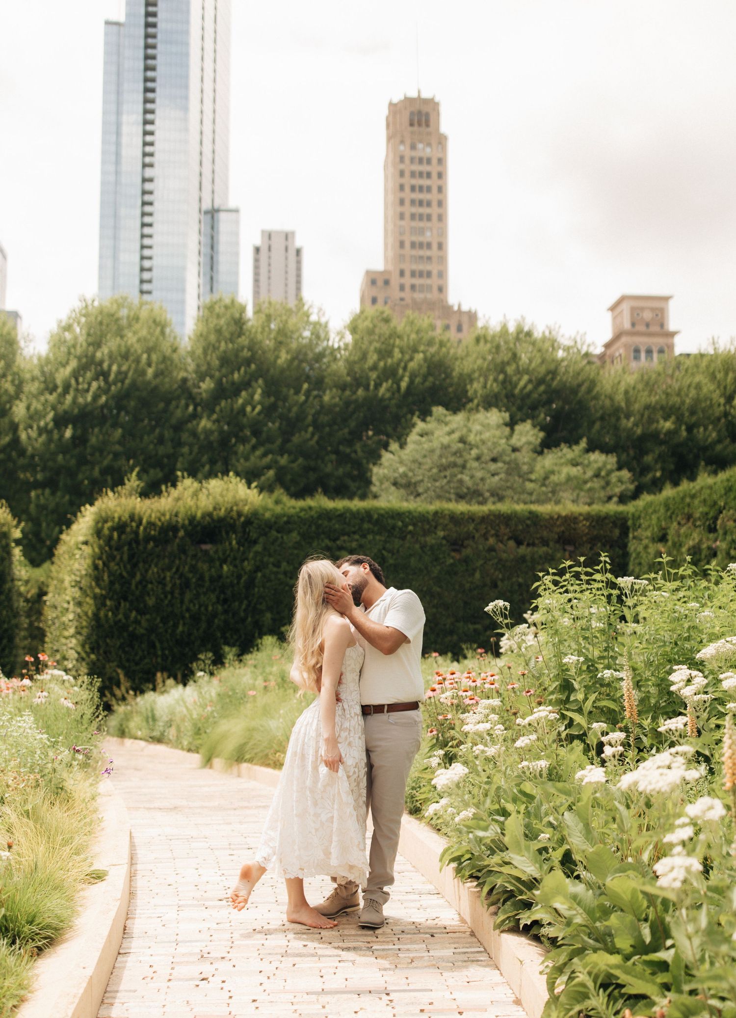 Couple sharing a romantic kiss in a lush garden pathway with city skyscrapers visible in the background.