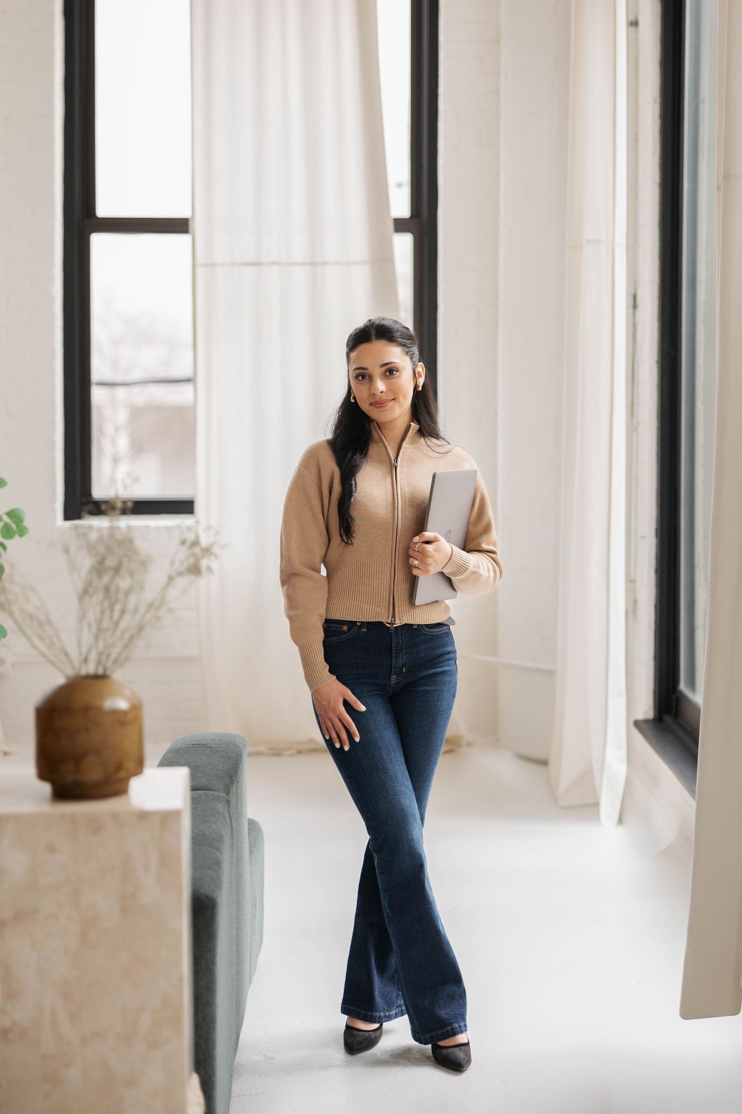Professional woman in beige jacket and flare jeans holding laptop, standing in modern bright office space.