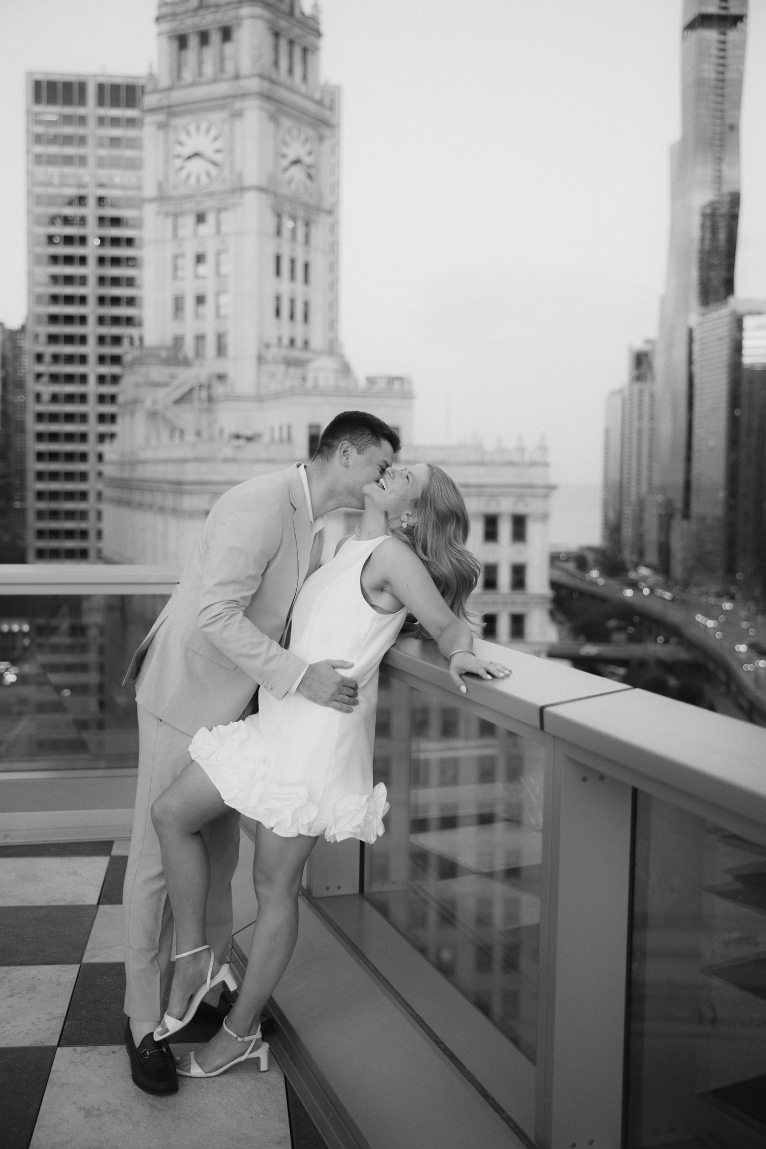 Black and white photo of couple kissing on rooftop with Chicago clock tower skyline in background.