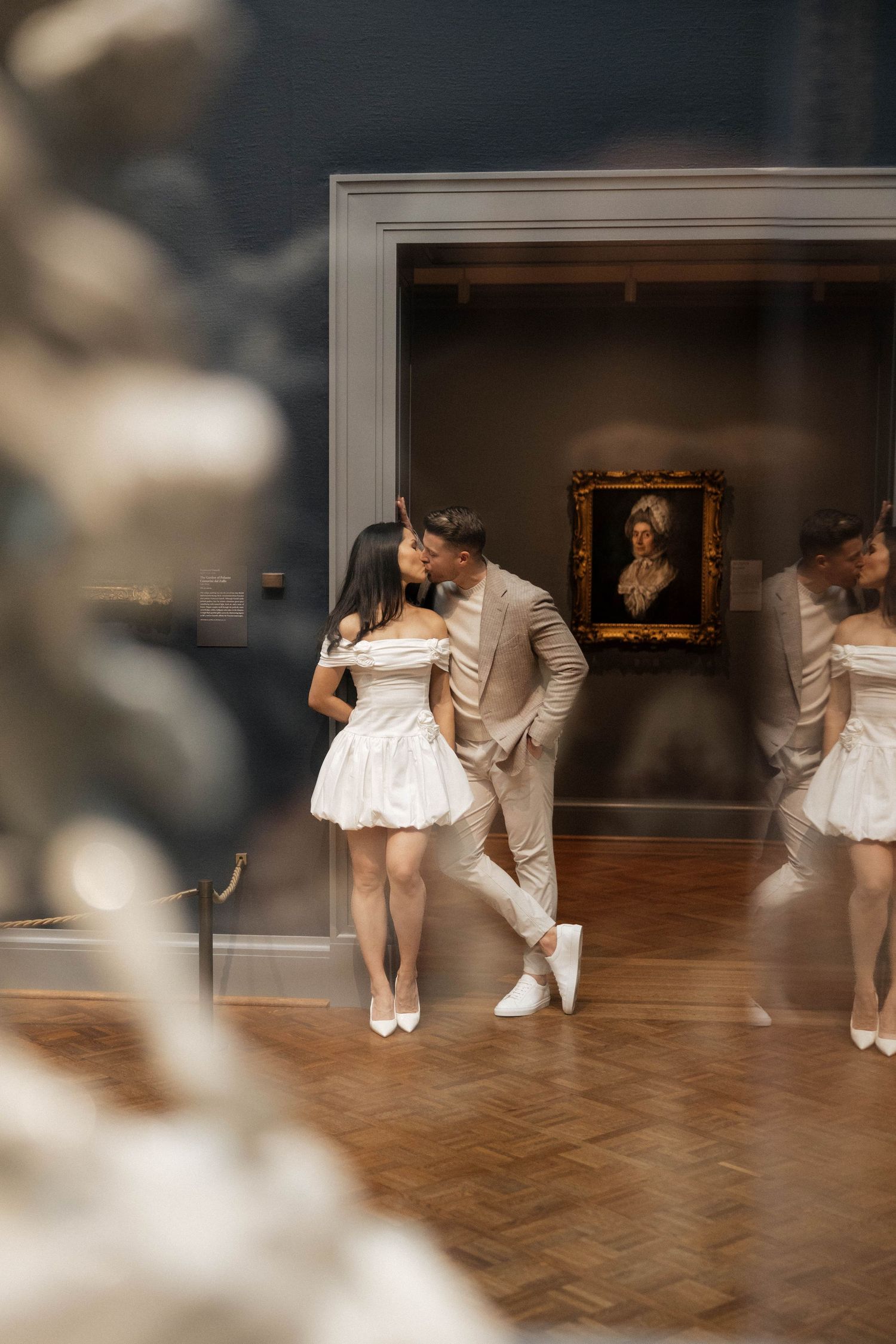 Couple in wedding attire sharing a romantic moment in an art museum gallery with a portrait painting in the background.