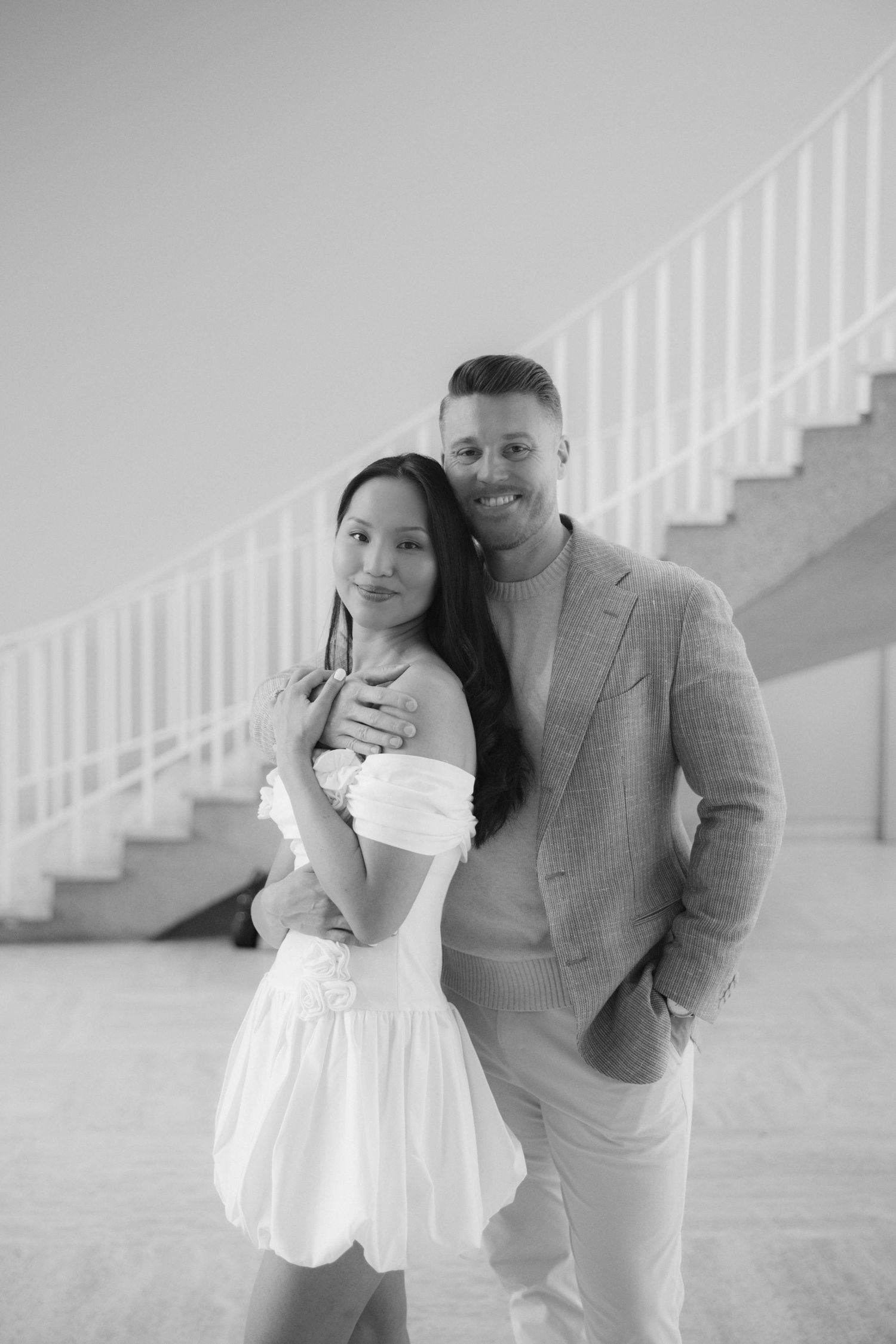 Black and white portrait of a smiling couple embracing in front of a spiral staircase.