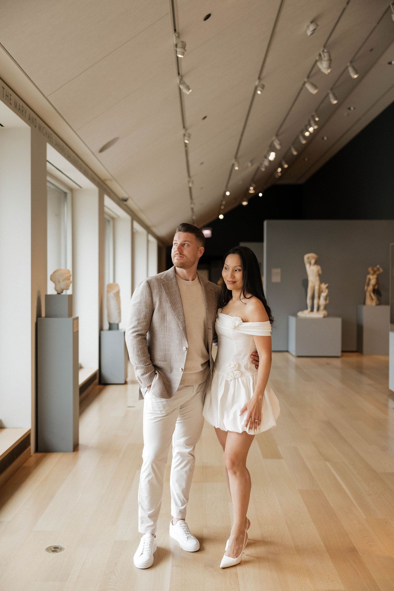 Elegant couple posing in a museum gallery with classical sculptures, man in grey blazer, woman in white mini dress.