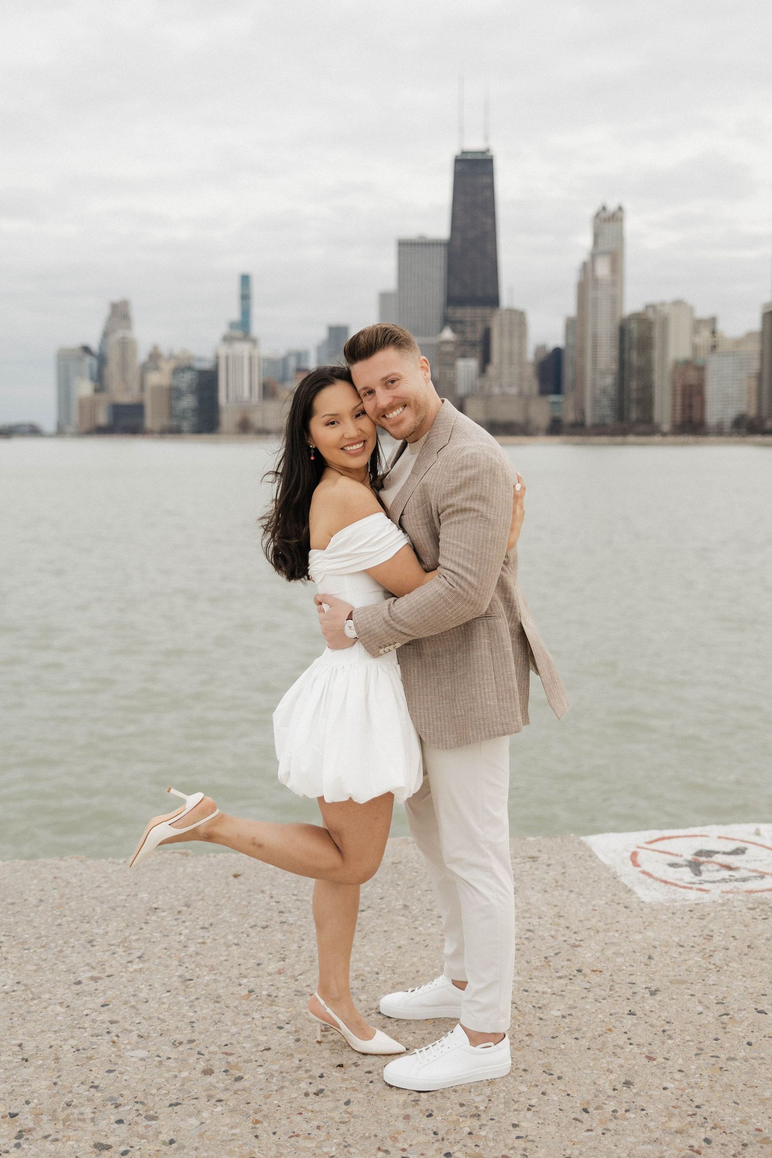 Couple embracing and smiling on Chicago lakefront with city skyline backdrop, woman in white dress, man in beige suit.