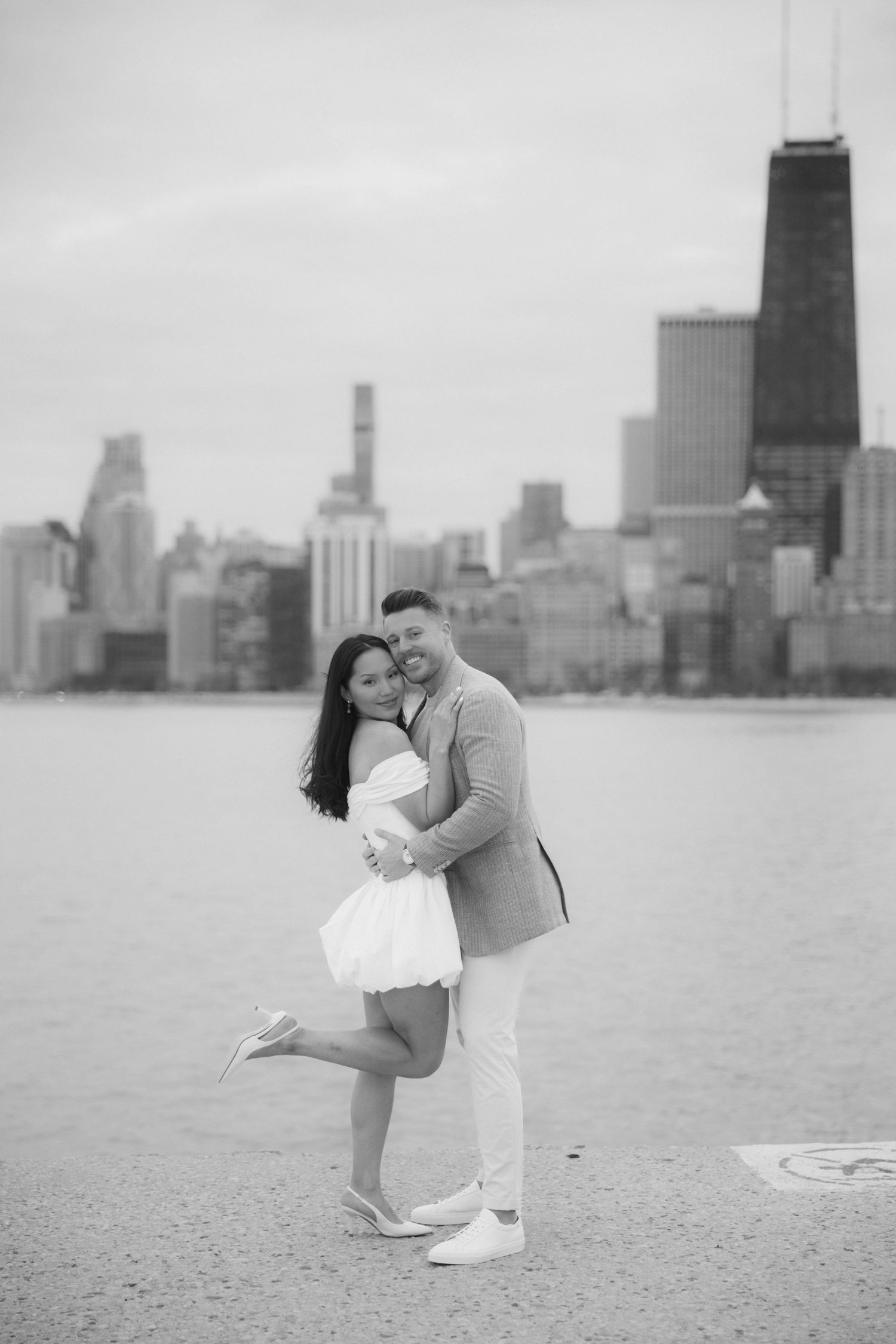 Black and white photo of couple hugging by Chicago skyline near the lakefront.