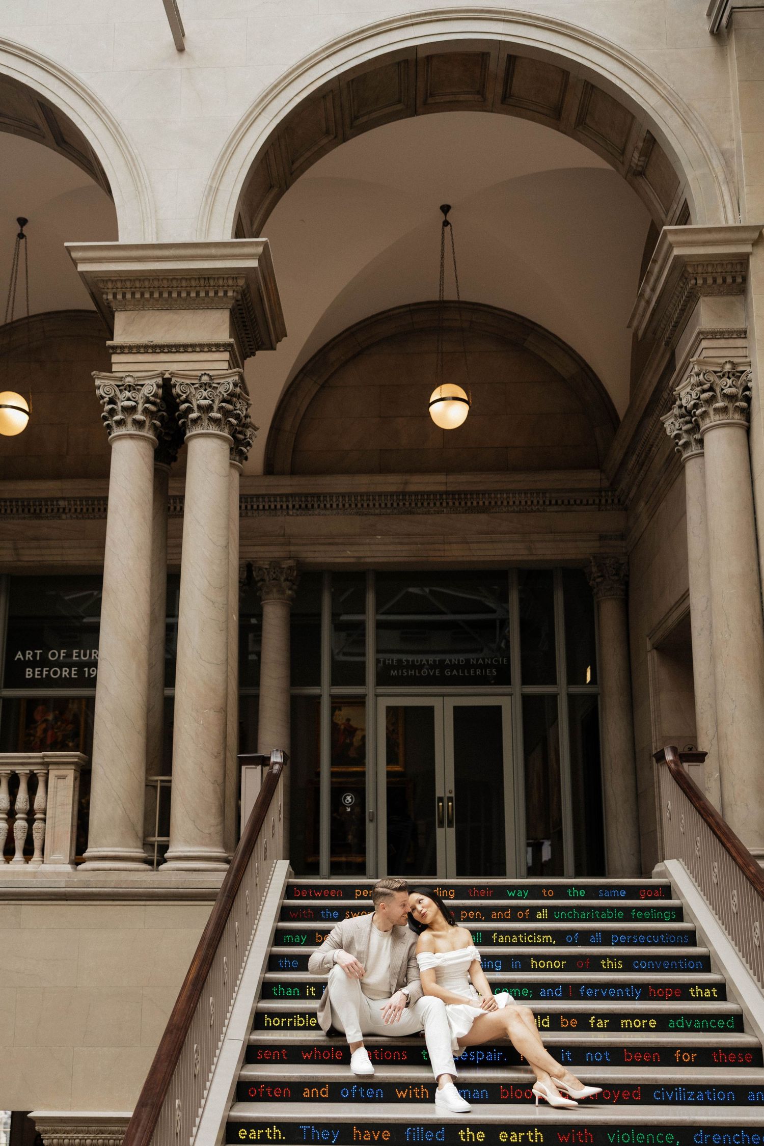 Couple sitting on colorful text-decorated steps outside a classical building with arched columns.