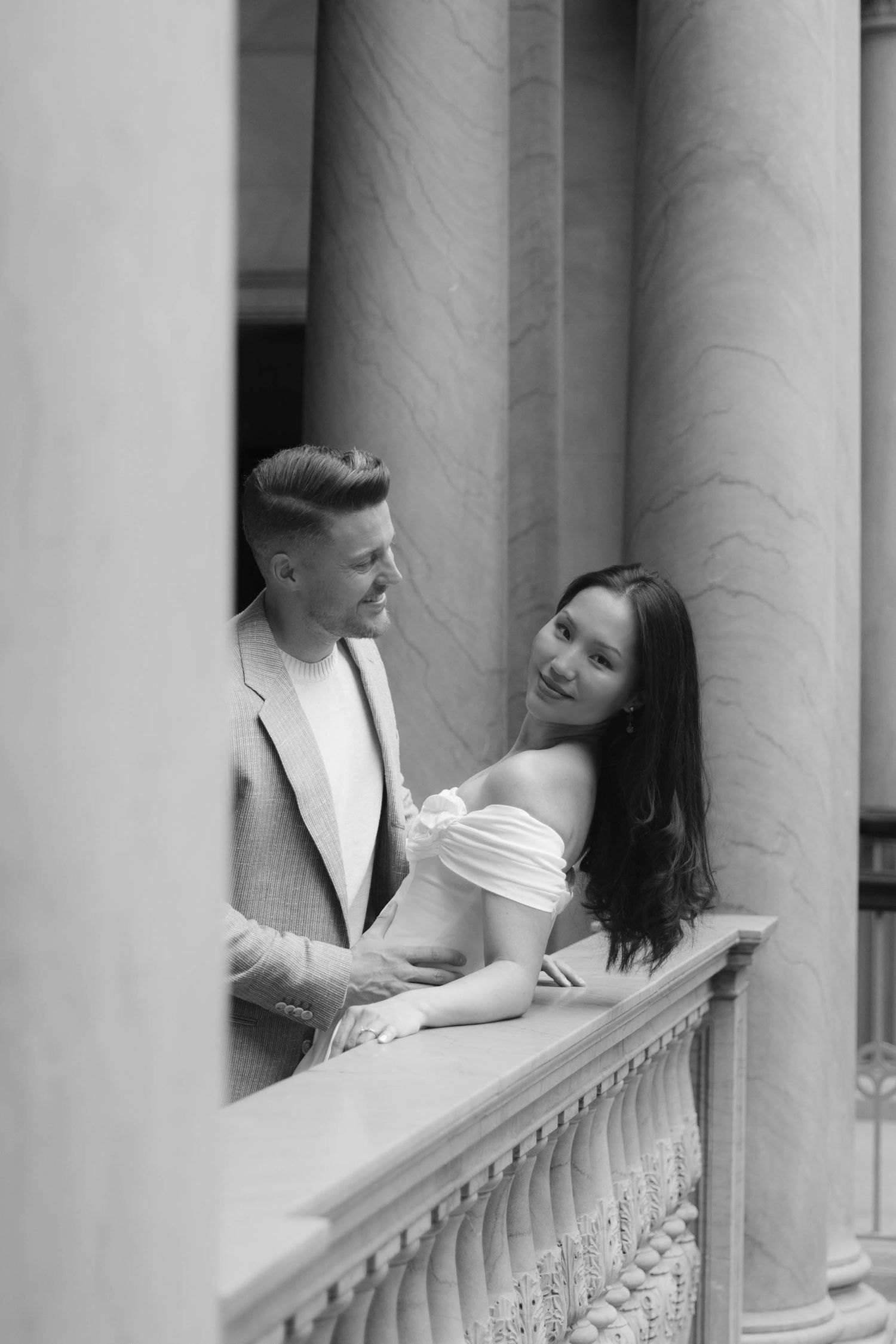 Black and white photo of a smiling couple standing between elegant columns on a balcony.