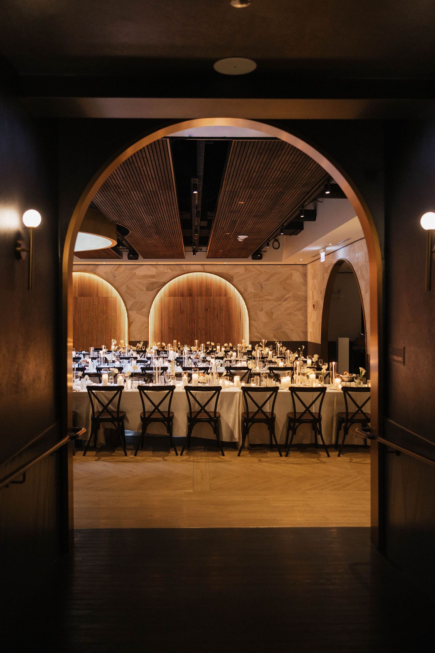 Elegant banquet hall viewed through arched doorway with long dining tables, warm lighting, and arched wall accents.