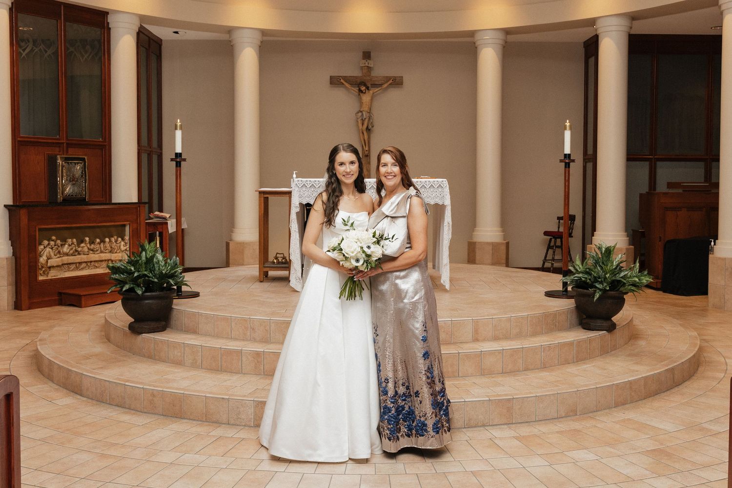 Bride in white gown and guest in blue floral dress pose together at church altar with crucifix backdrop.