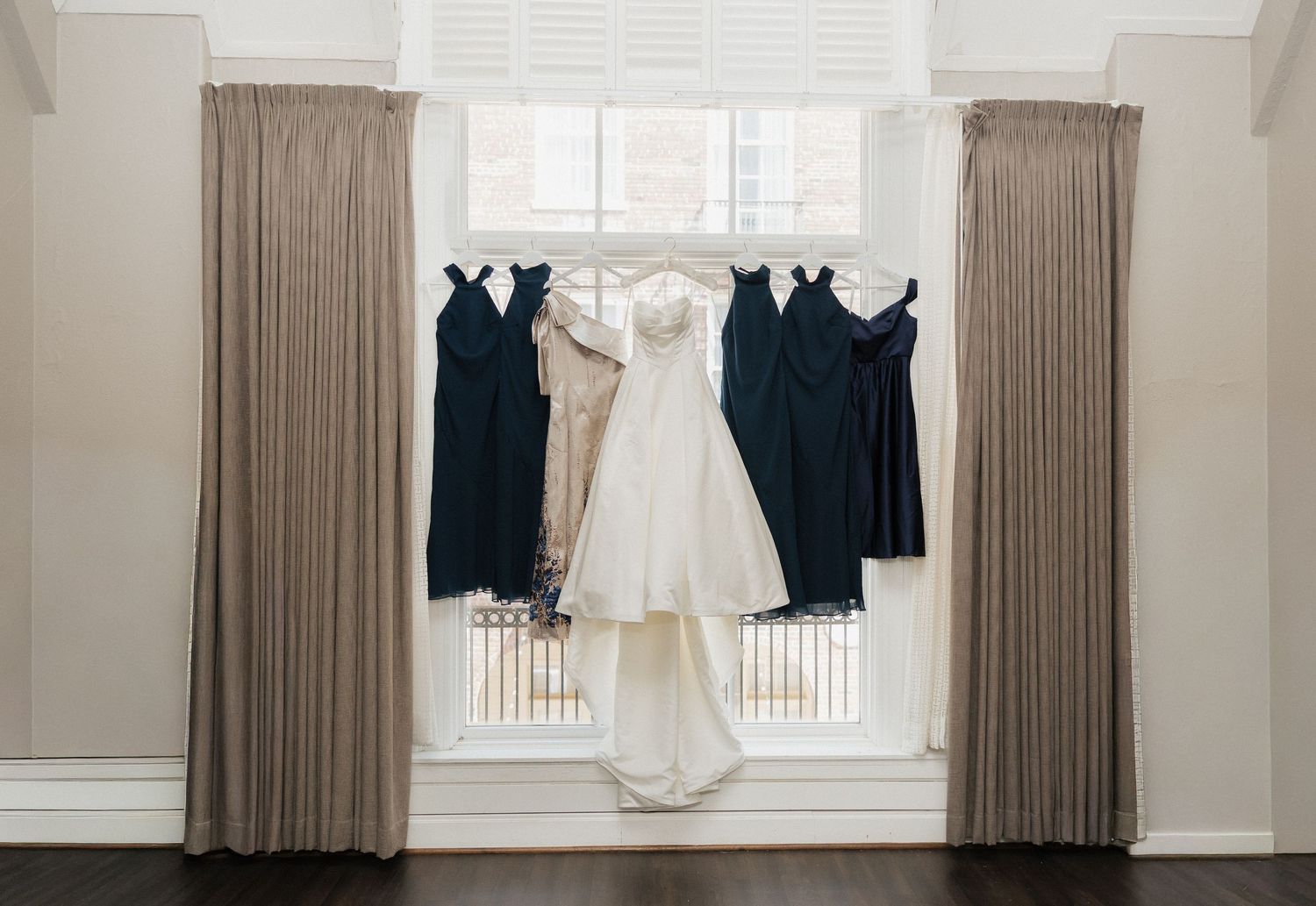 White wedding gown and navy bridesmaid dresses hanging in a sunlit window flanked by elegant taupe curtains.