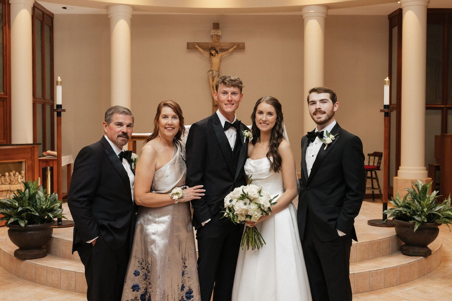 Wedding party of five posing at church altar with crucifix, bride in white gown holding bouquet, groom and guests in formal attire.