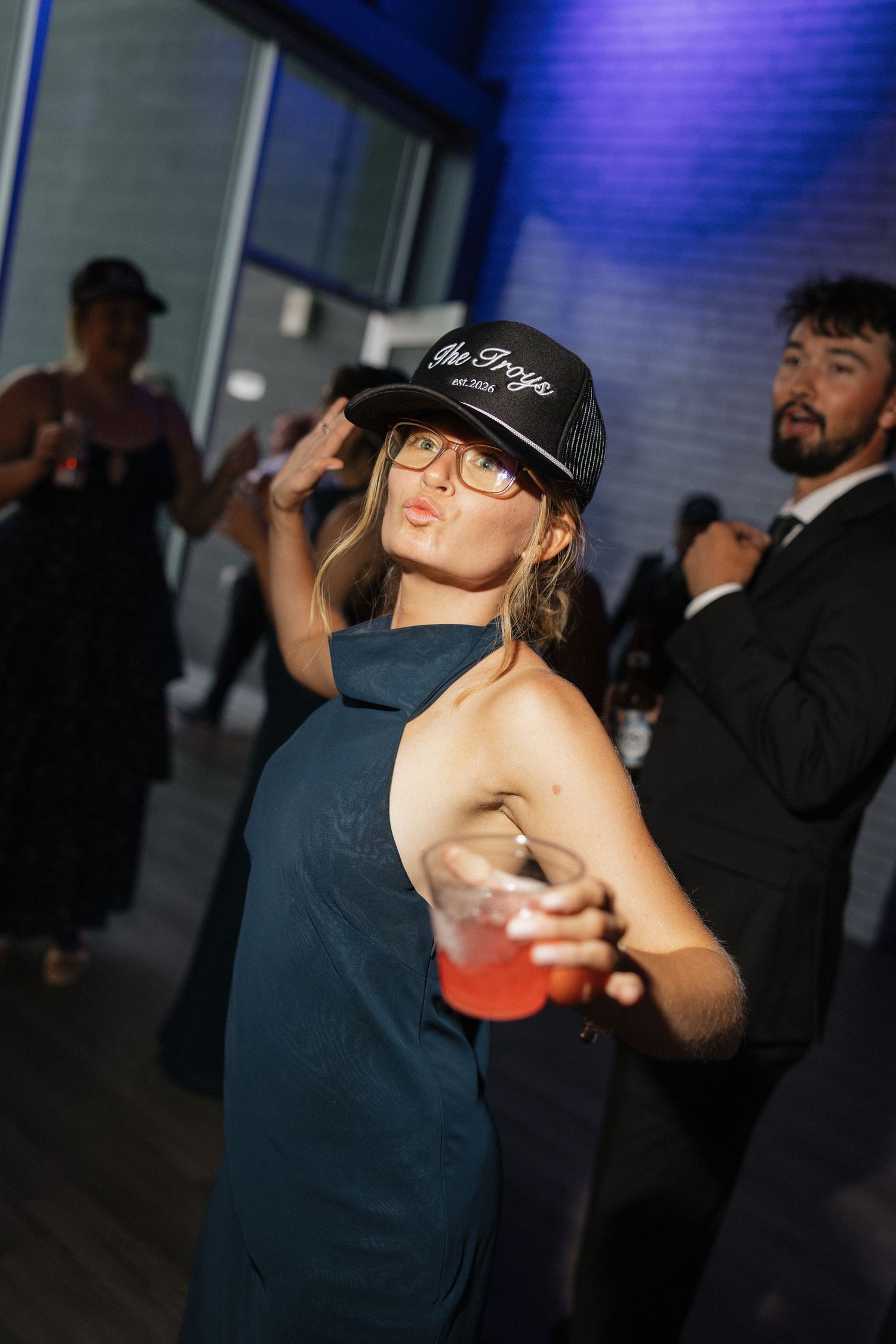 Woman in teal dress and black trucker hat poses with pink drink at elegant evening party.