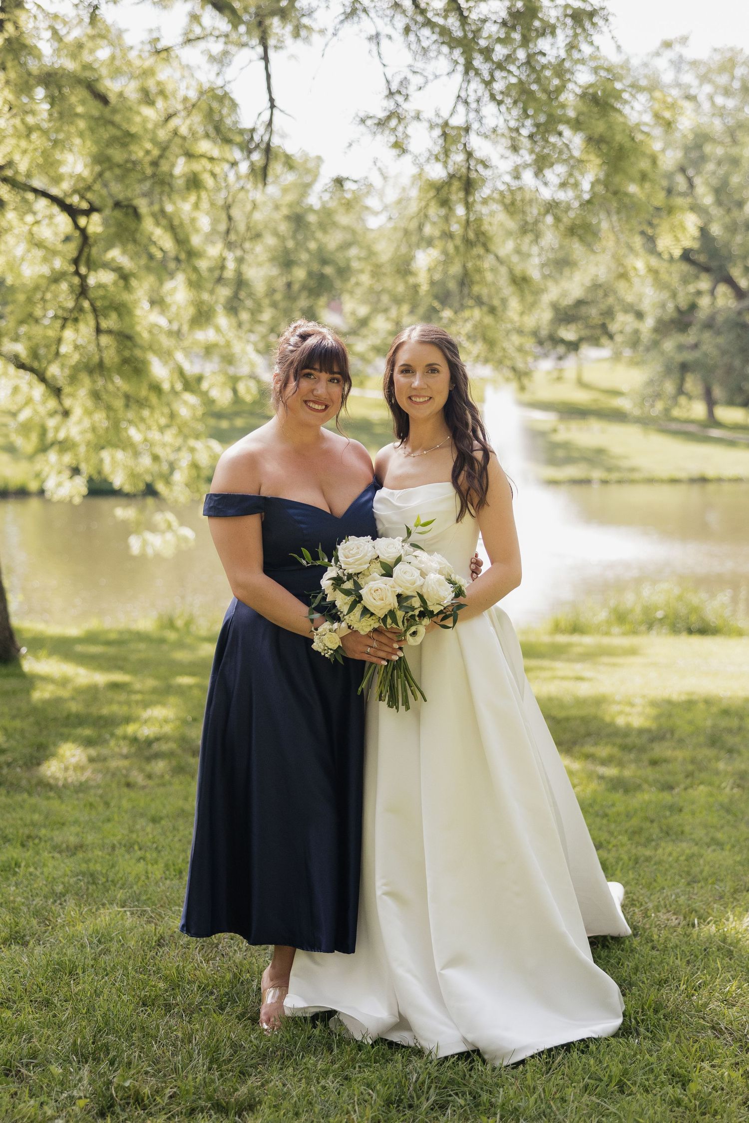Bride in white gown and bridesmaid in navy off-shoulder dress pose with bouquet in lush park setting.