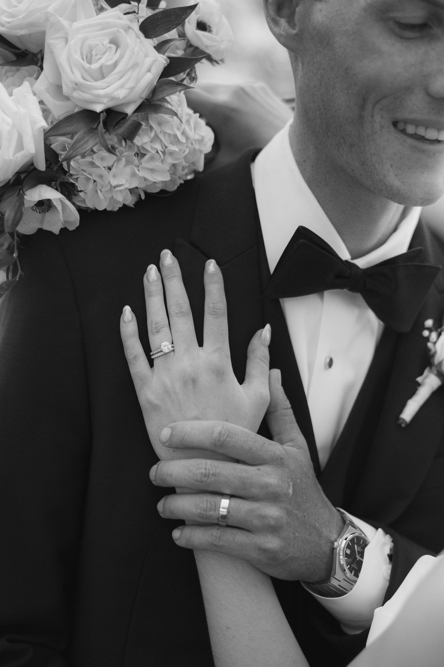 Black and white photo of newlyweds showing wedding rings, groom in tuxedo with bow tie, bride holding white rose bouquet.