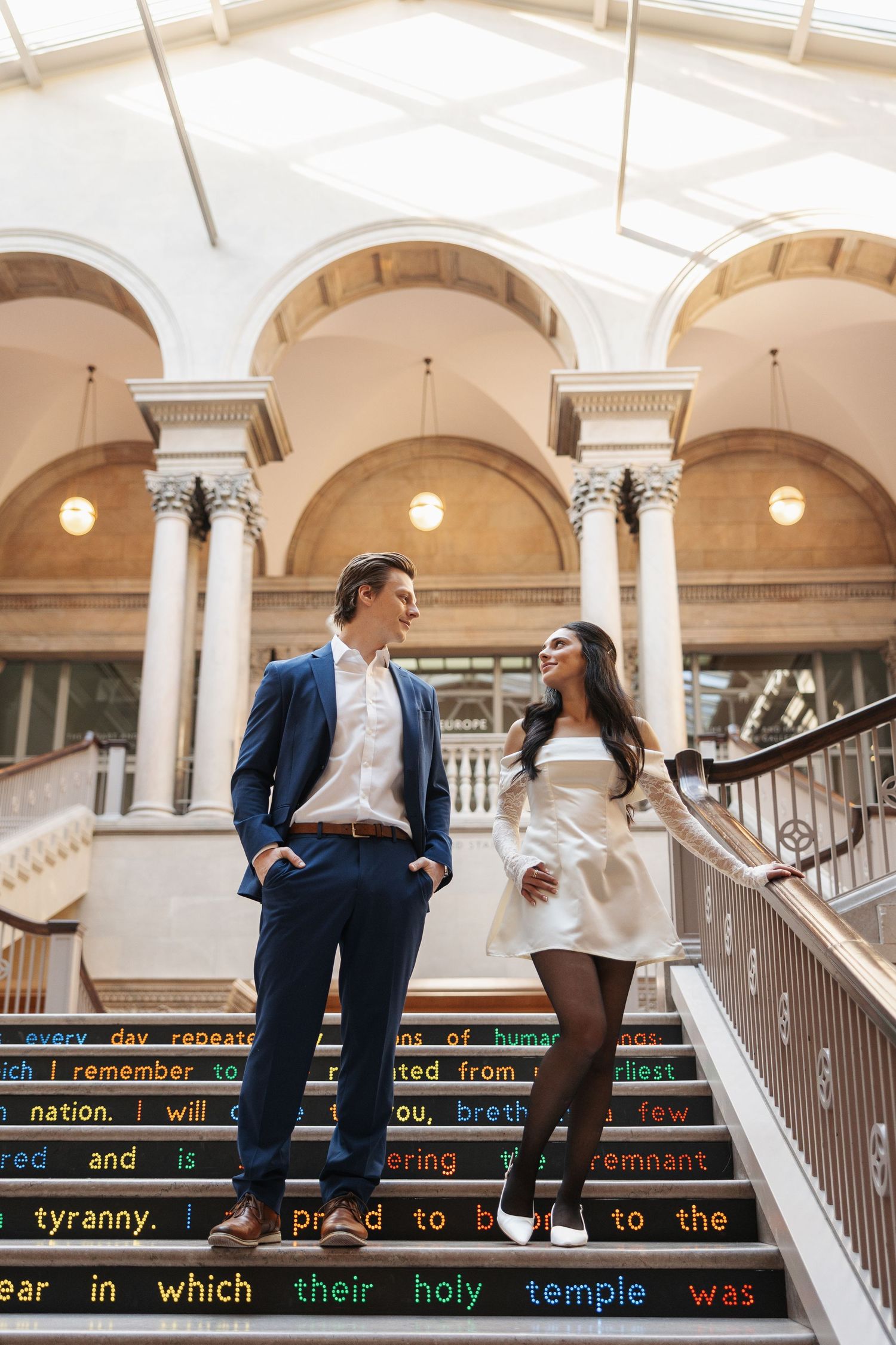 A couple sits on marble steps with illuminated text displays visible in the background.