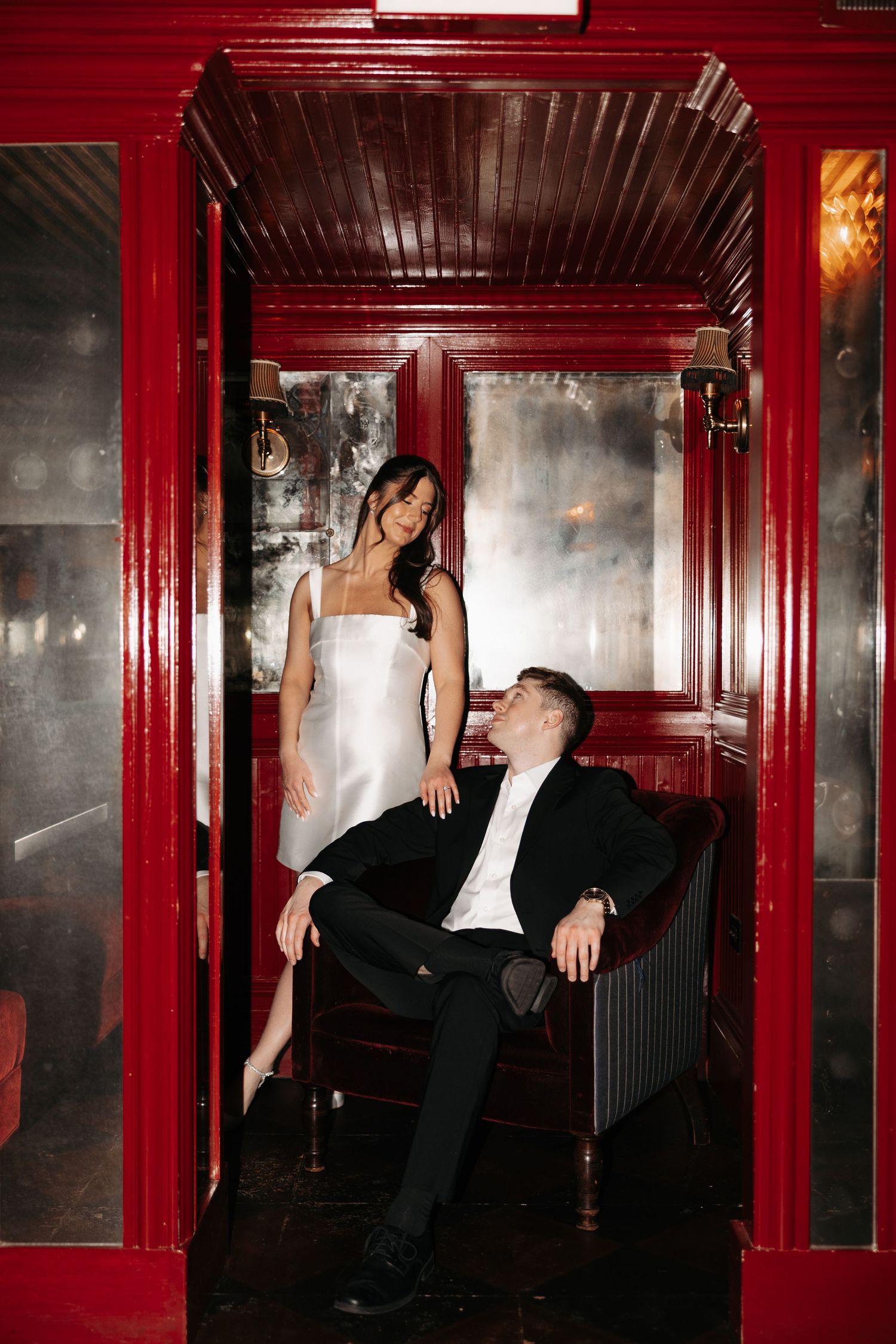 A couple poses in an elegant red-framed elevator, with a woman in white and a man in black formal attire.